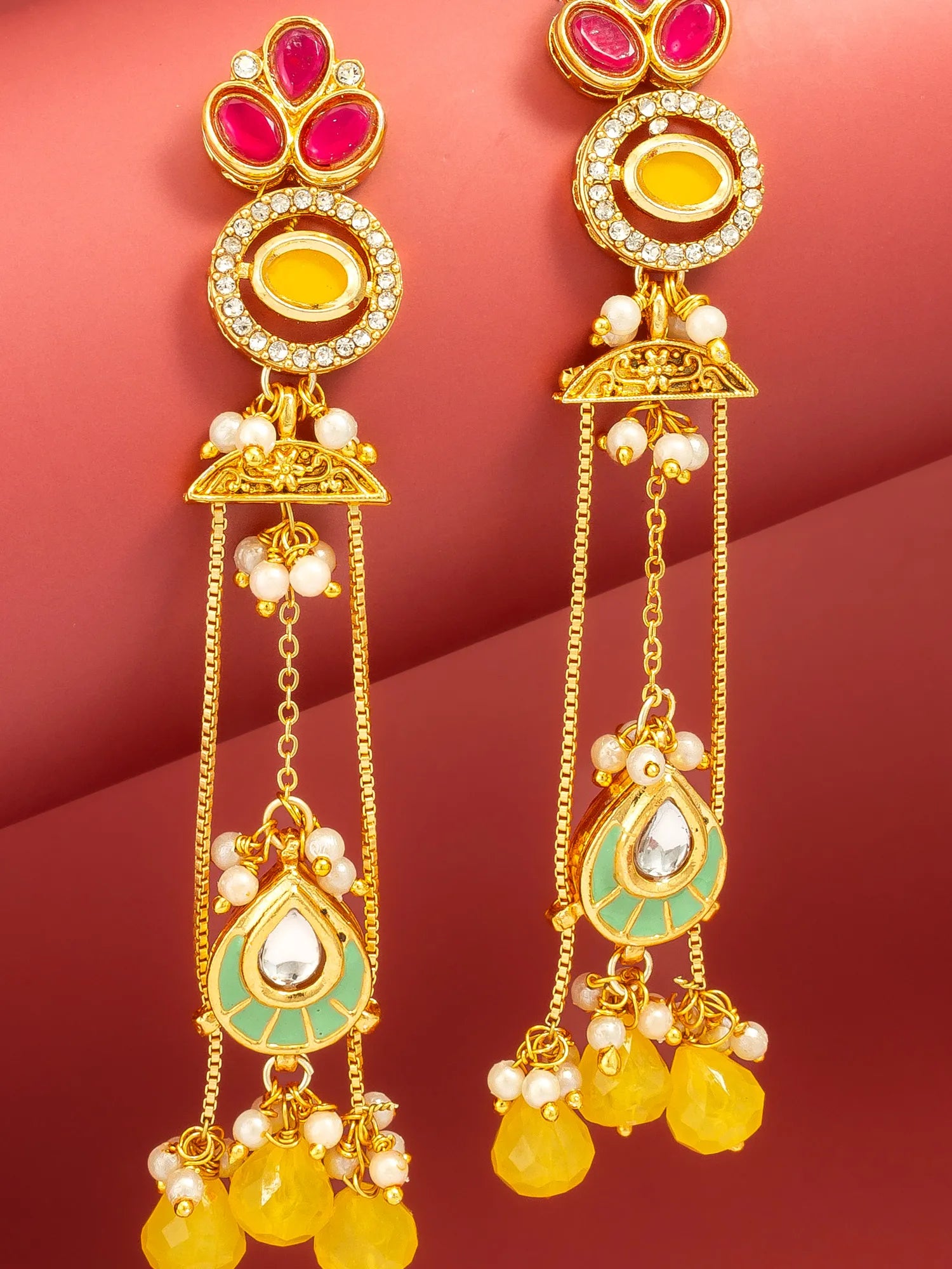 A pair of gold-plated Indian jhumka earrings decorated with pink and yellow stones, white faux pearls, and light green enamel, displayed against a solid dark red background.