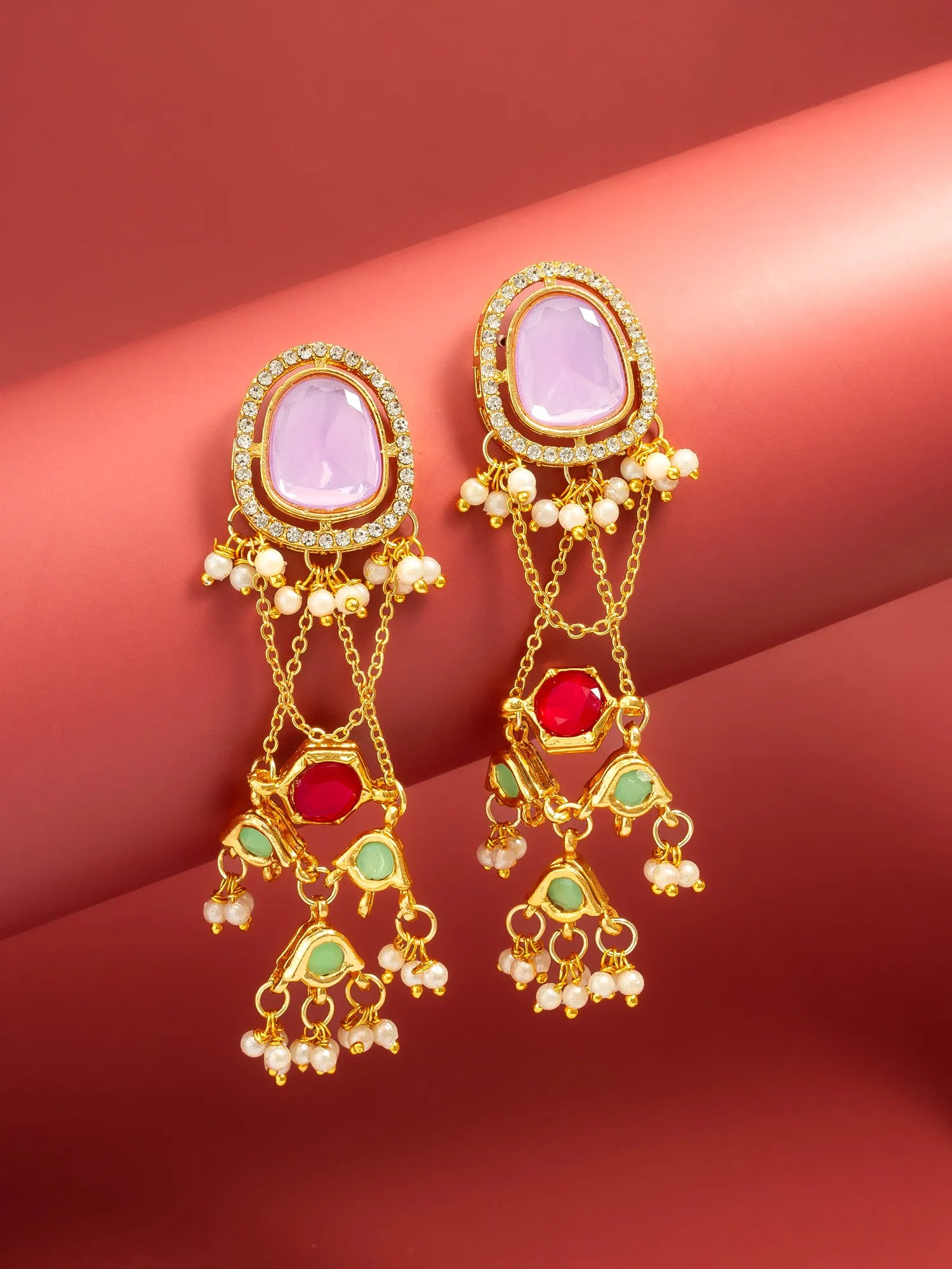 A pair of ornate gold-toned dangle earrings with oval light purple stones, red stones, green stones, and small white bead accents, displayed on a pinkish-red curved surface.