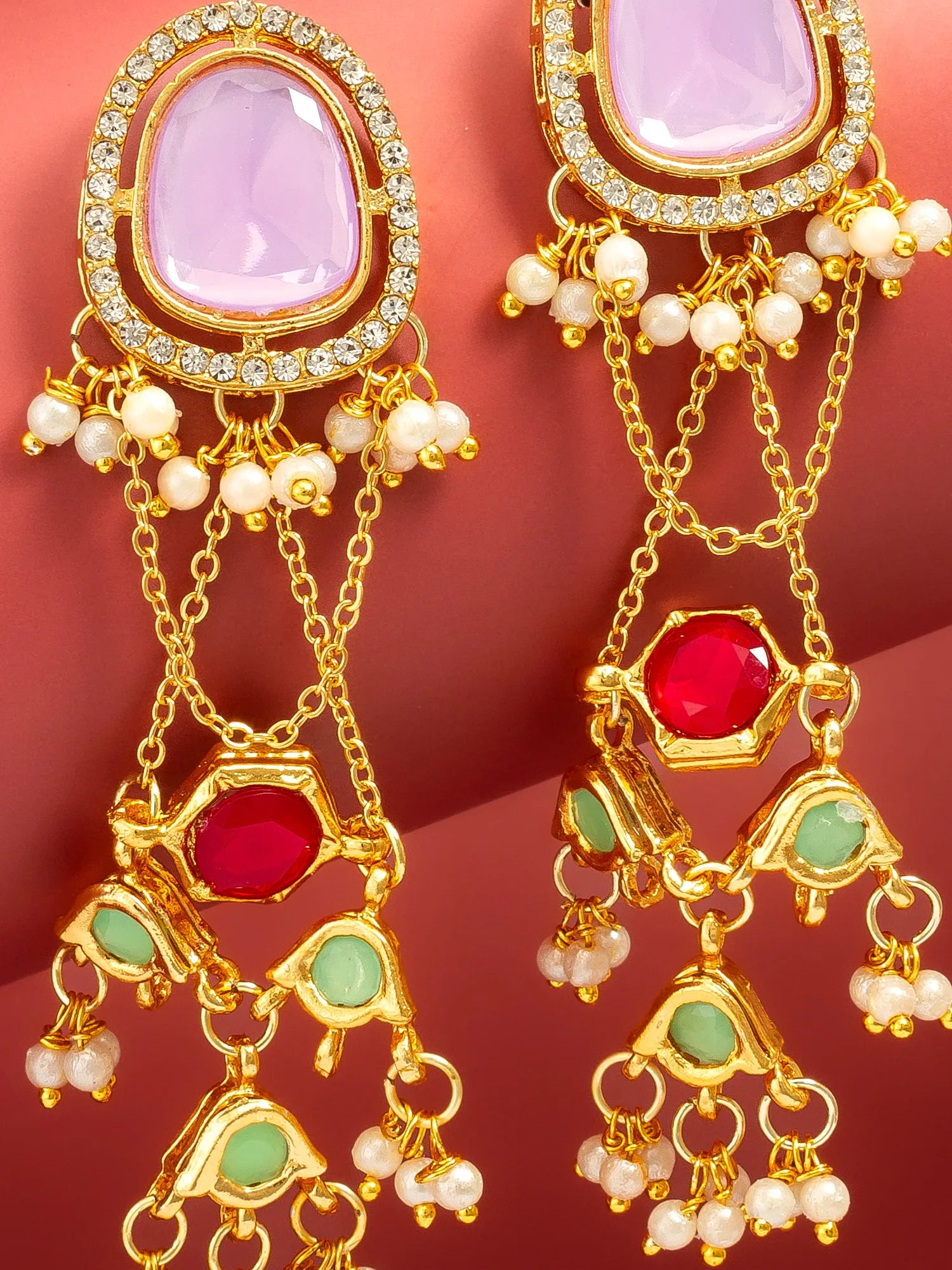 A pair of ornate gold-toned dangle earrings with large pale pink stones at the top, clear rhinestone borders, white pearl-like beads, and red and light green stone accents, shown against a warm pink background.