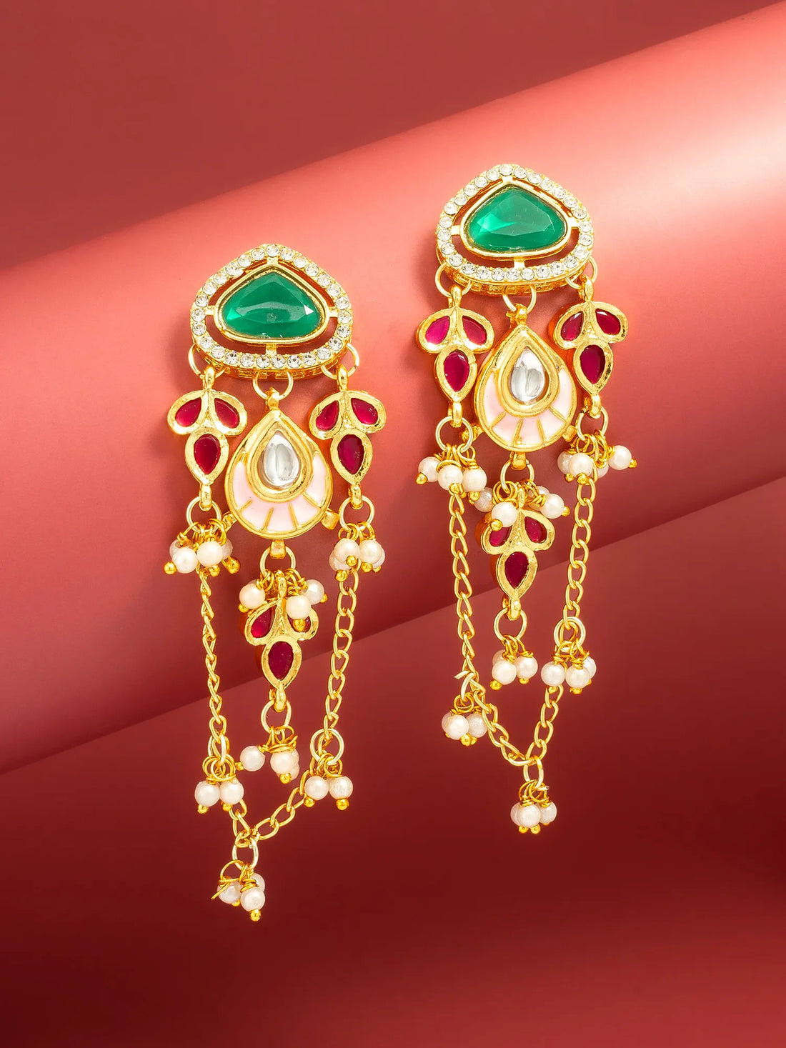 A pair of ornate gold-toned dangle earrings with green teardrop-shaped stones, white pearl-like beads, and small red gemstone accents, displayed against a warm red background.