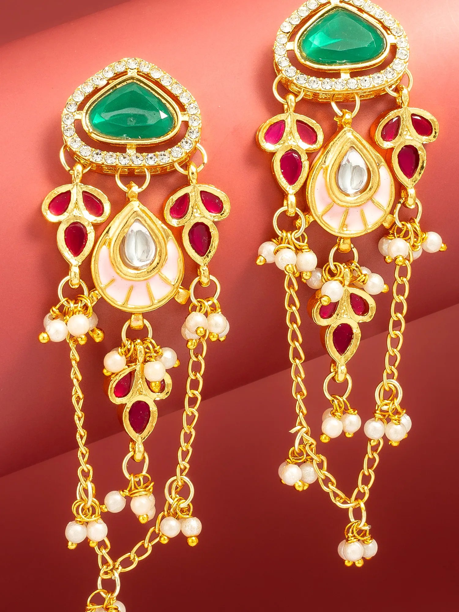 A pair of ornate gold-toned dangle earrings with green teardrop-shaped stones, pink-red oval accents, white pearl-like beads, and clear rhinestone detailing, shown on a warm red background.