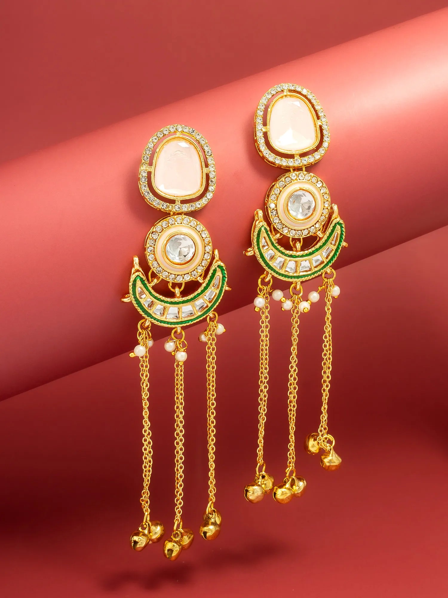 A pair of gold-toned traditional Indian dangle earrings with white stones, green enamel, and small pearl-like beads on a red background.