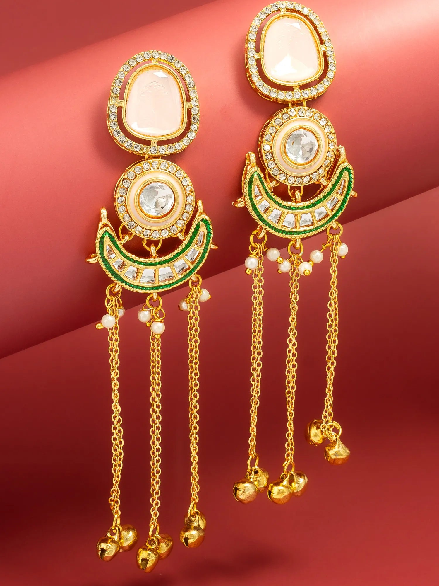 A pair of gold-plated Indian jhumka earrings with white stone accents, green enamel detailing, and dangling chain bells, displayed on a curved red background.