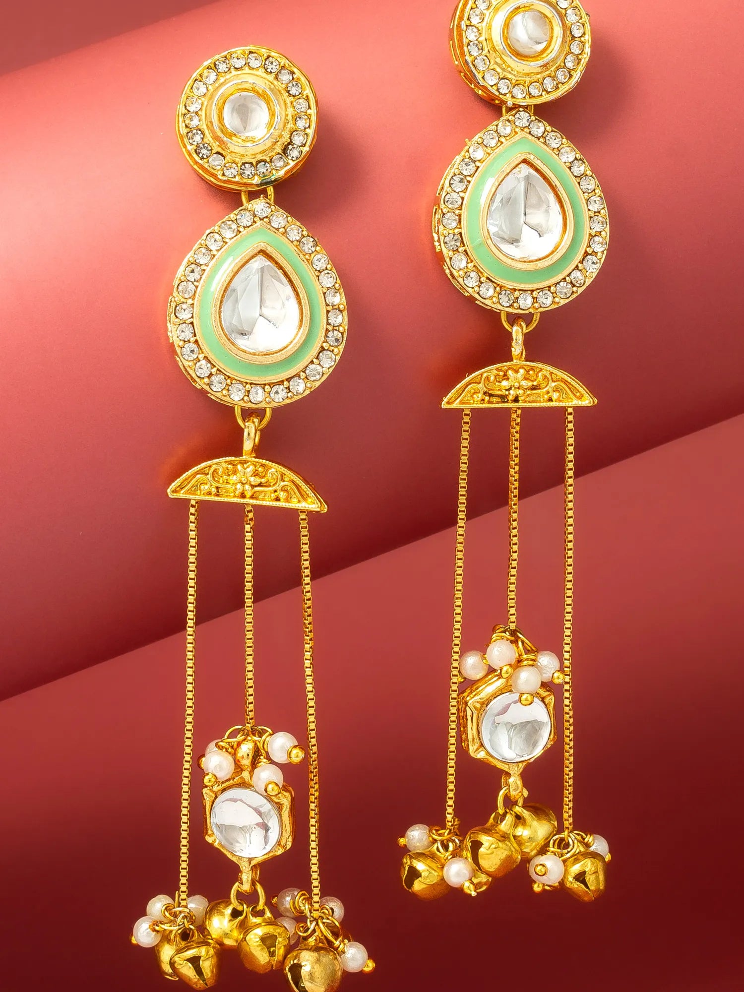 A pair of gold-toned traditional Indian Kundan dangle earrings with light green enamel, clear crystals, and small pearl-like beads, displayed on a textured red background.