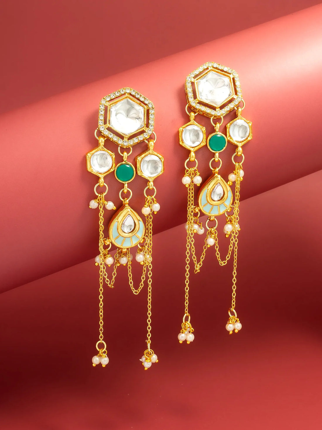 A pair of gold-toned dangle earrings with white stones, green stones, and small pearl-like beads, displayed against a pinkish-red background.