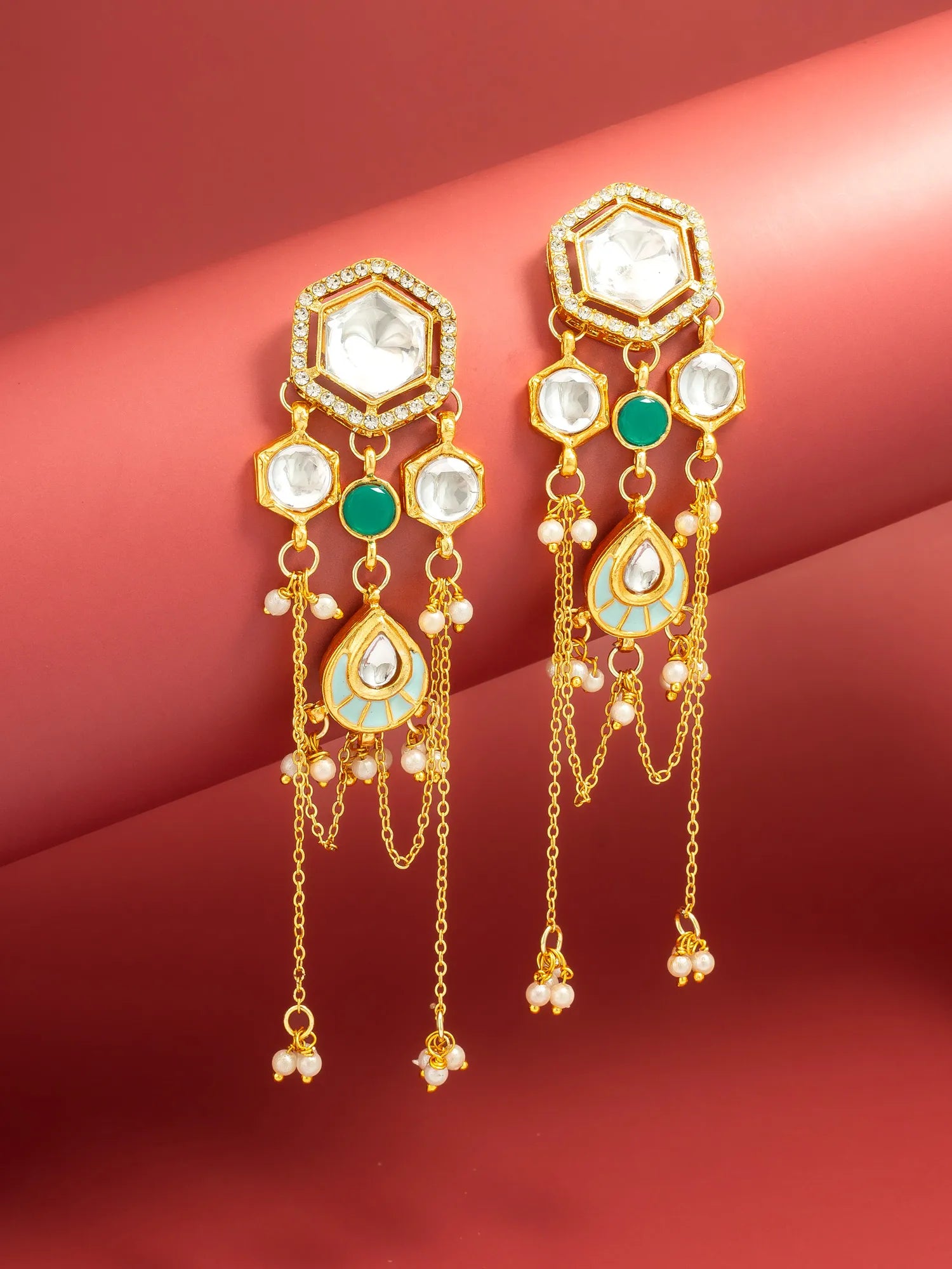A pair of gold-toned dangle earrings with white stones, green stones, and small pearl-like beads, displayed against a pinkish-red background.