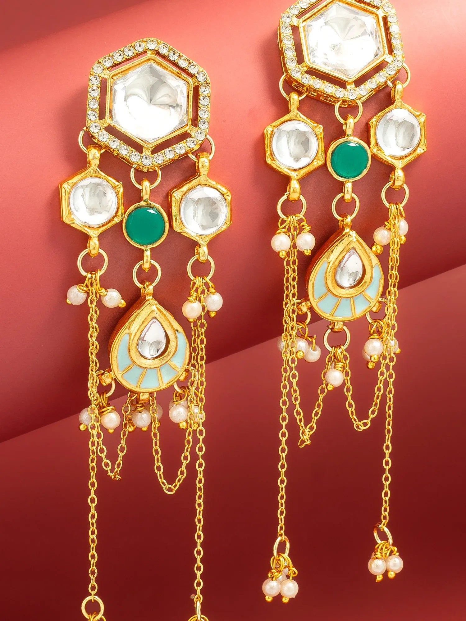A pair of gold-plated Indian style dangle earrings with white crystals, green stones, and light blue enamel details, shown against a dark red background.