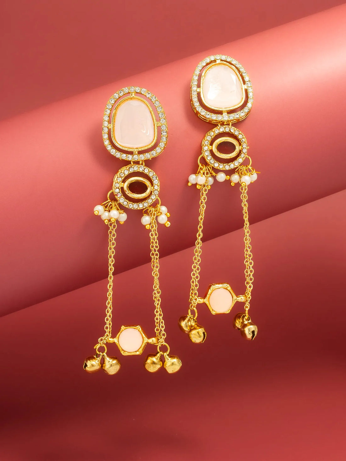 A pair of gold-toned statement earrings with oval white stone settings, small clear stones, and dangling chain elements with bell charms, displayed on a pinkish-red background.