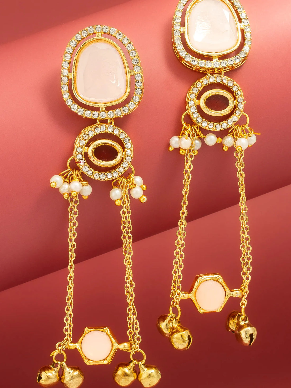 A pair of gold-toned statement earrings with oval white stone settings, small clear stones, and dangling chain elements with bell charms, displayed on a pinkish-red background.