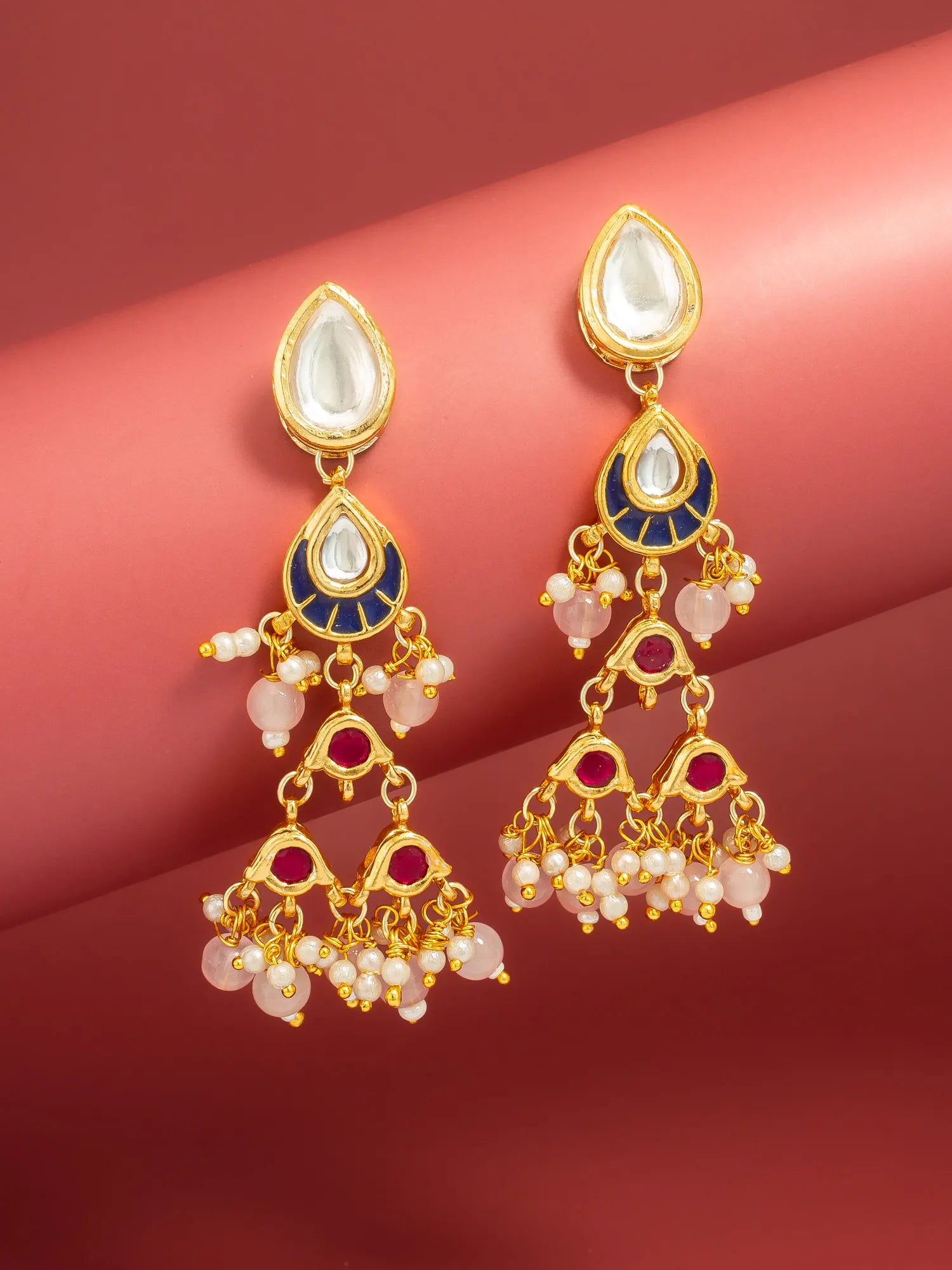 A pair of gold-plated dangle earrings with white stone tops, navy blue enamel sections, and pink pearl-like bead clusters, displayed on a curved pink background.
