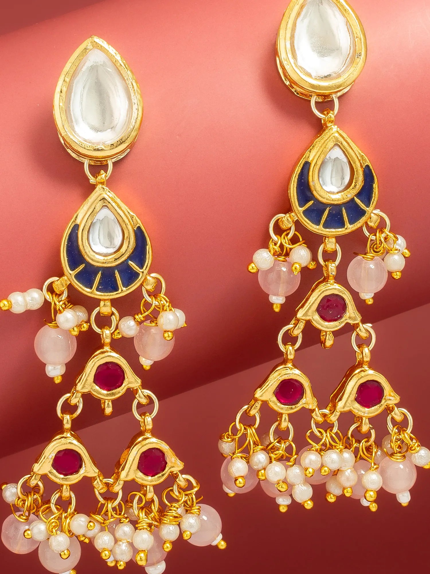 A pair of gold-plated Indian jhumka earrings with white stone studs, blue enamel, pink stone beads, and white pearl drops, set against a solid coral-pink background.