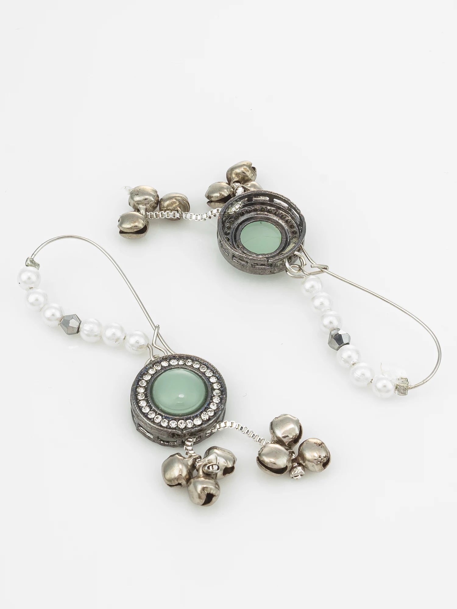 A pair of silver-toned dangle earrings with oval light green stone centers, surrounded by small clear stones, decorated with white pearl bead strands and dangling silver bell charms, shown on a plain white background.