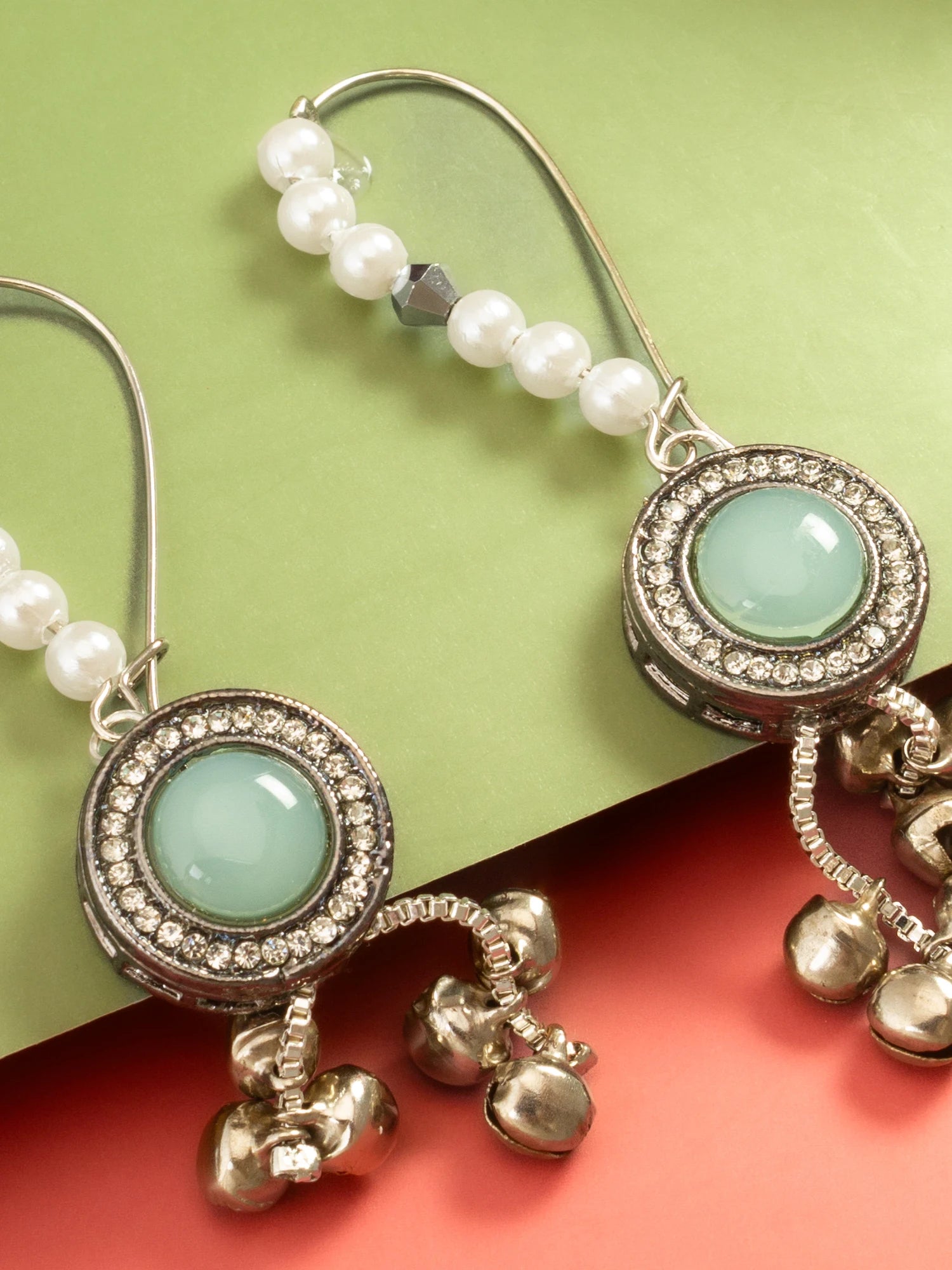 A pair of silver-toned dangle earrings with white pearl beads, a light green oval stone in a rhinestone-set oval frame, and dangling silver bells, shown on a split green and coral background.