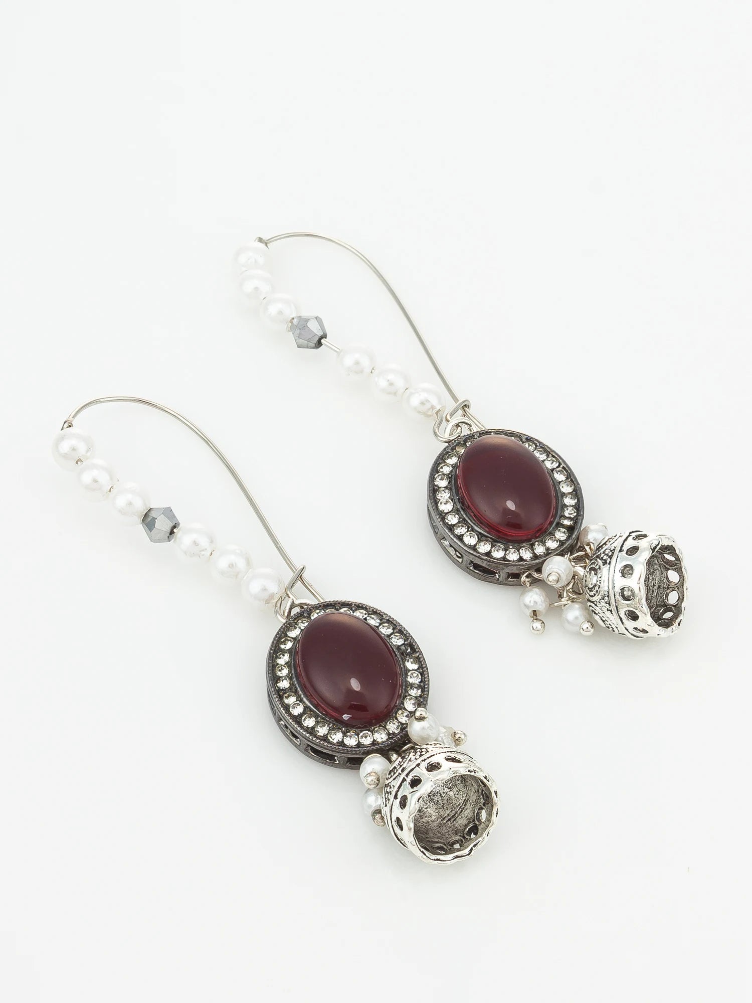 A pair of silver-toned dangle earrings featuring oval dark red cabochon stones surrounded by small clear rhinestones, with white pearl bead strands and dangling silver bell charms, shown on a plain white background.