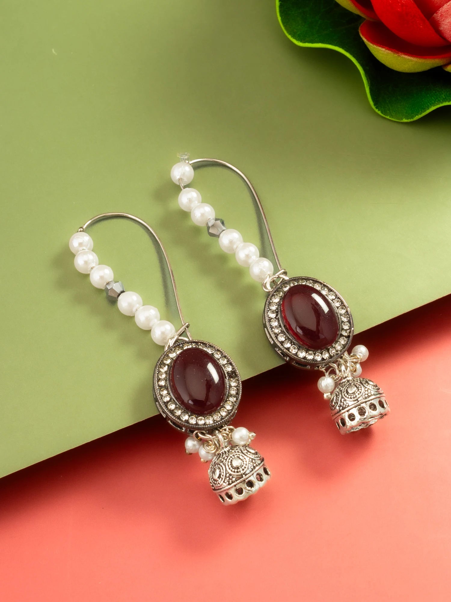 A pair of silver-toned Indian jhumka earrings with oval dark red gemstones, small white pearl beads, and ornate bell-shaped dangling charms, displayed on a split green and coral background.