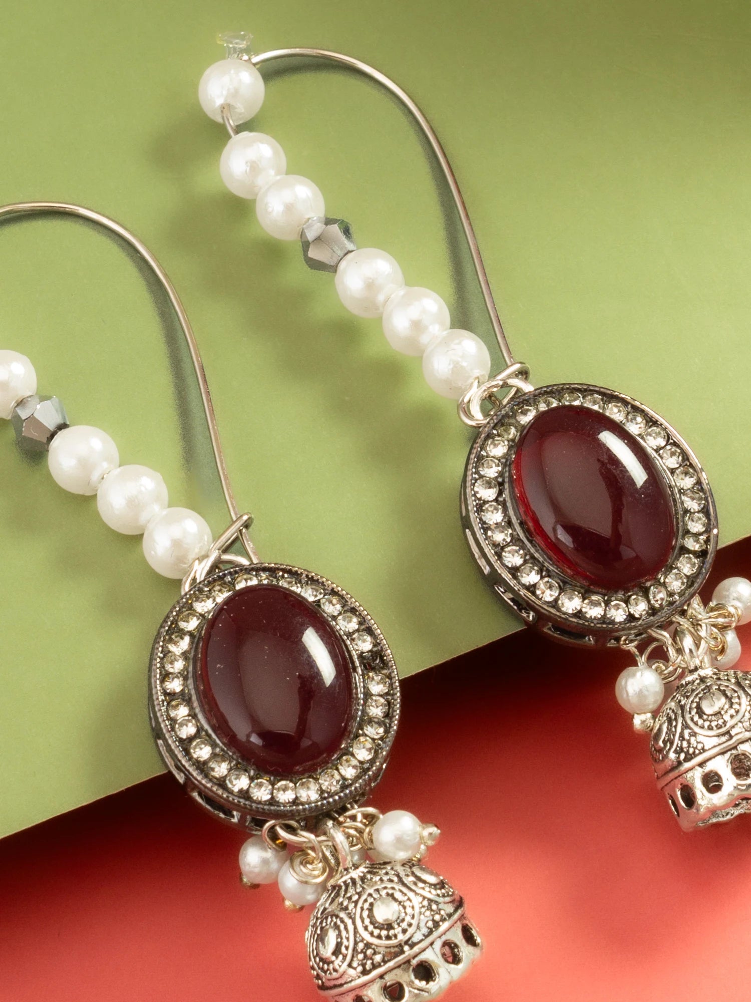 A pair of traditional Indian jhumka earrings with white pearl beads, silver-toned filigree borders, and oval deep red cabochon stones, displayed on a split green and red background.