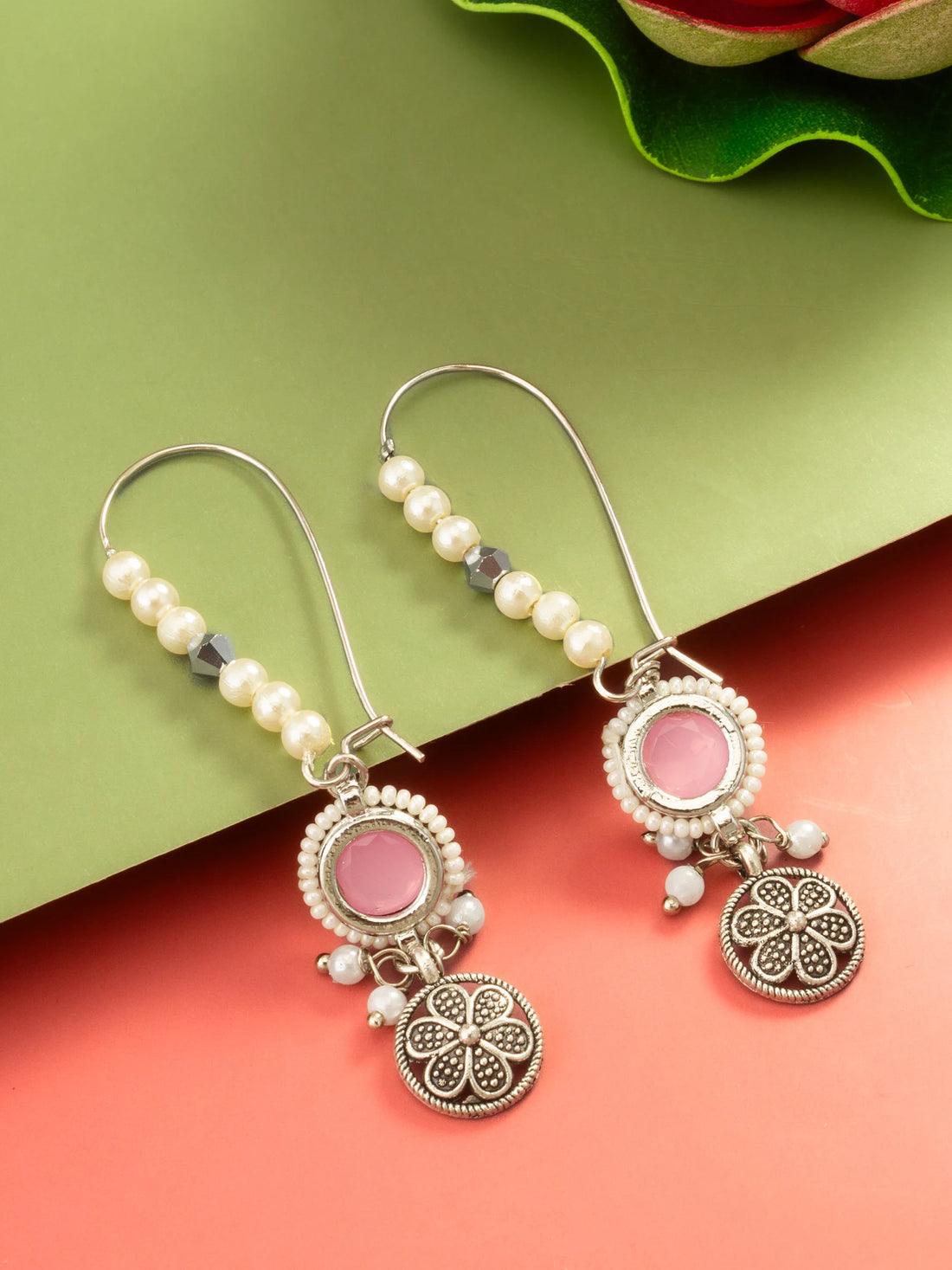 A pair of bohemian-style dangle earrings with silver-toned filigree, white faux pearls, and pink stone accents, displayed on a split green and coral background.
