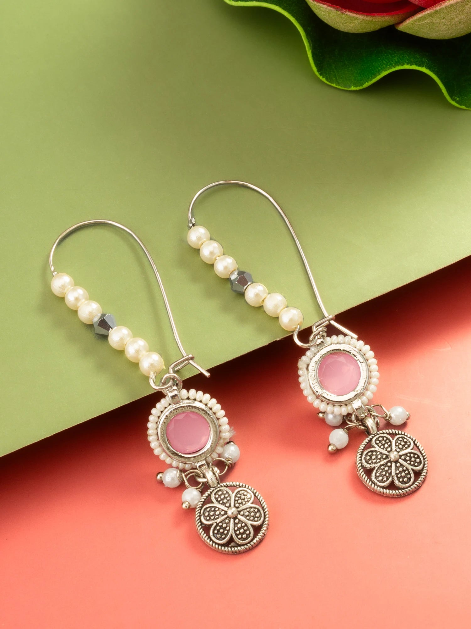 A pair of bohemian-style dangle earrings with silver-toned filigree, white faux pearls, and pink stone accents, displayed on a split green and coral background.