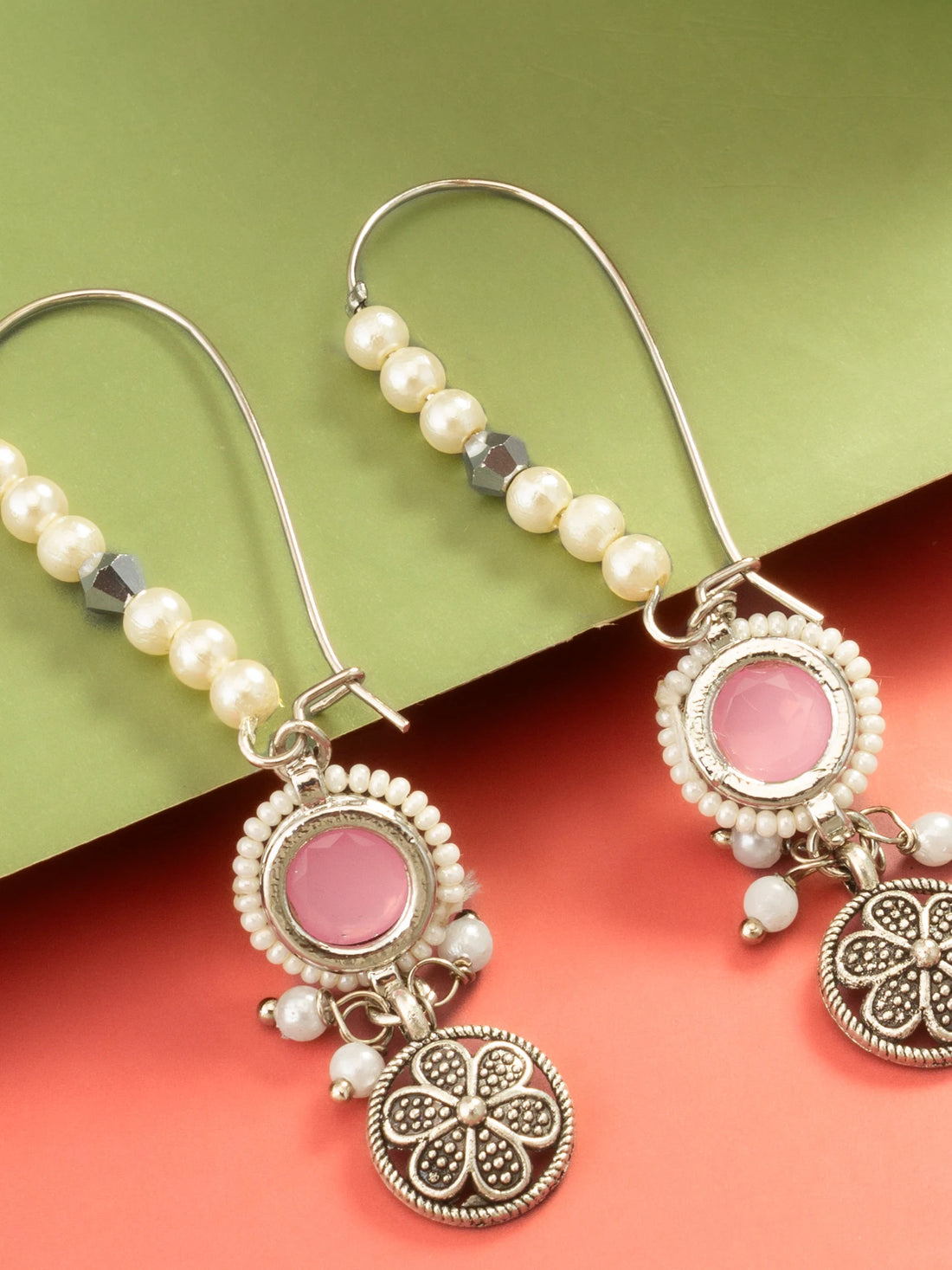 A pair of bohemian-style dangle earrings with silver-toned filigree, white faux pearls, and pink stone accents, displayed on a split green and coral background.
