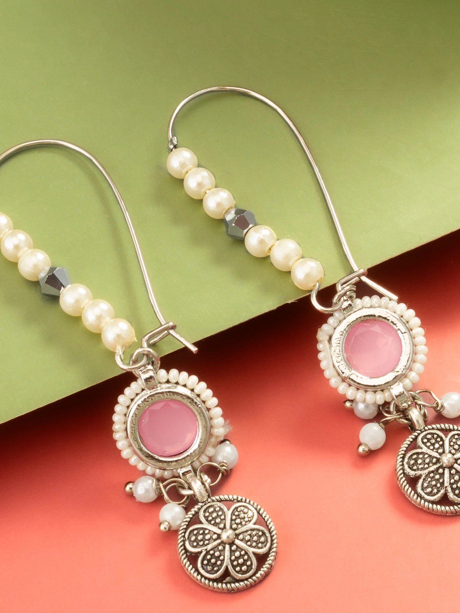 A pair of silver-toned dangle earrings with white pearl beads, a round pink stone, and a dangling ornate silver charm, displayed on a split green and coral background.