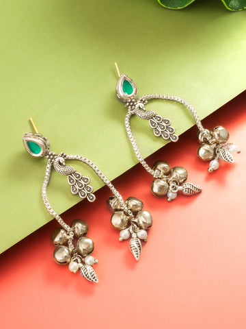 A pair of silver-toned dangle earrings with green teardrop-shaped stones, small clear stones, and dangling floral and leaf charms, arranged on a split green and coral background.