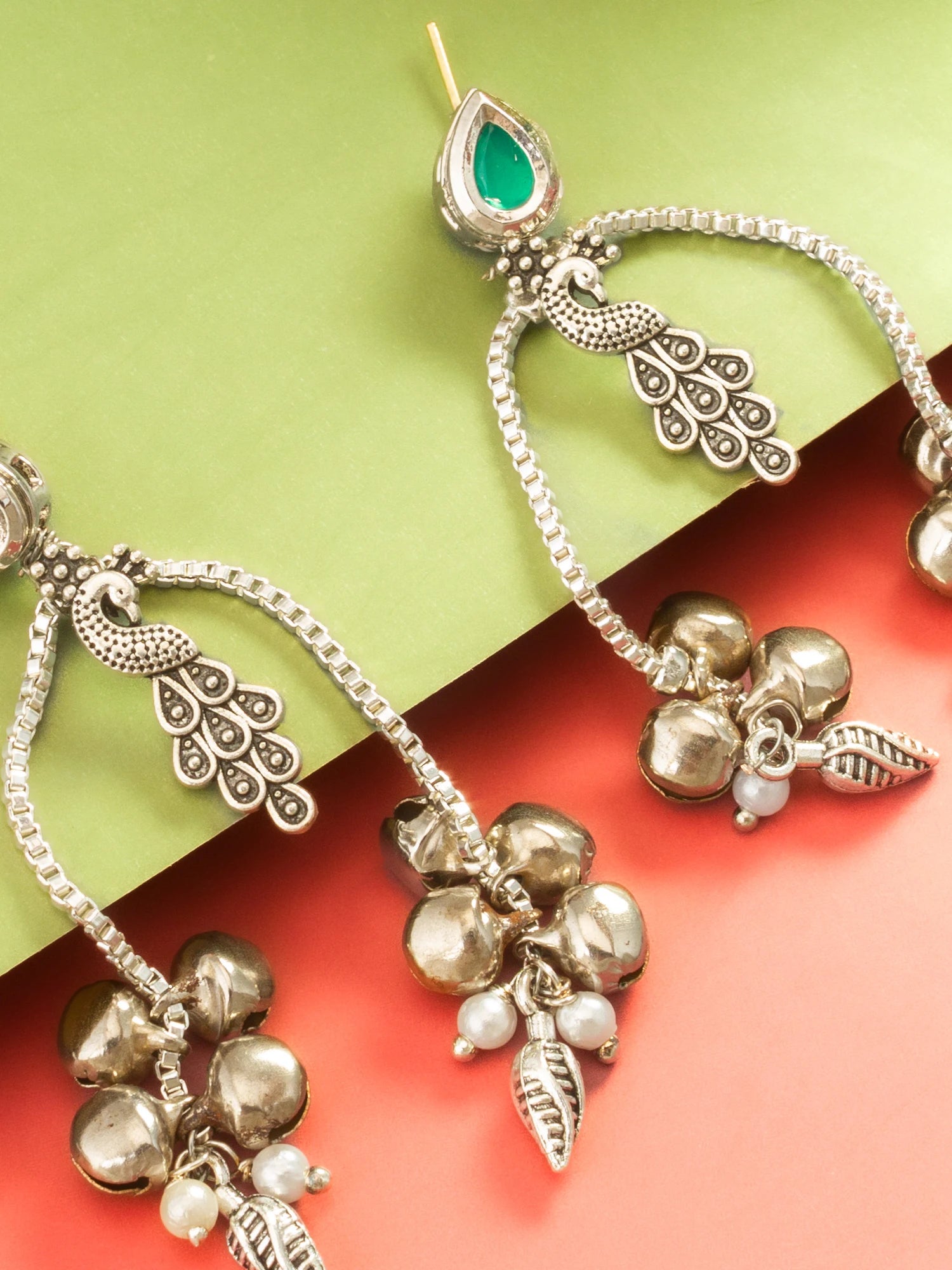 Silver-toned Indian jhumka earrings with peacock motifs, a green teardrop stone, and dangling bell charms, styled on a split green and coral background.