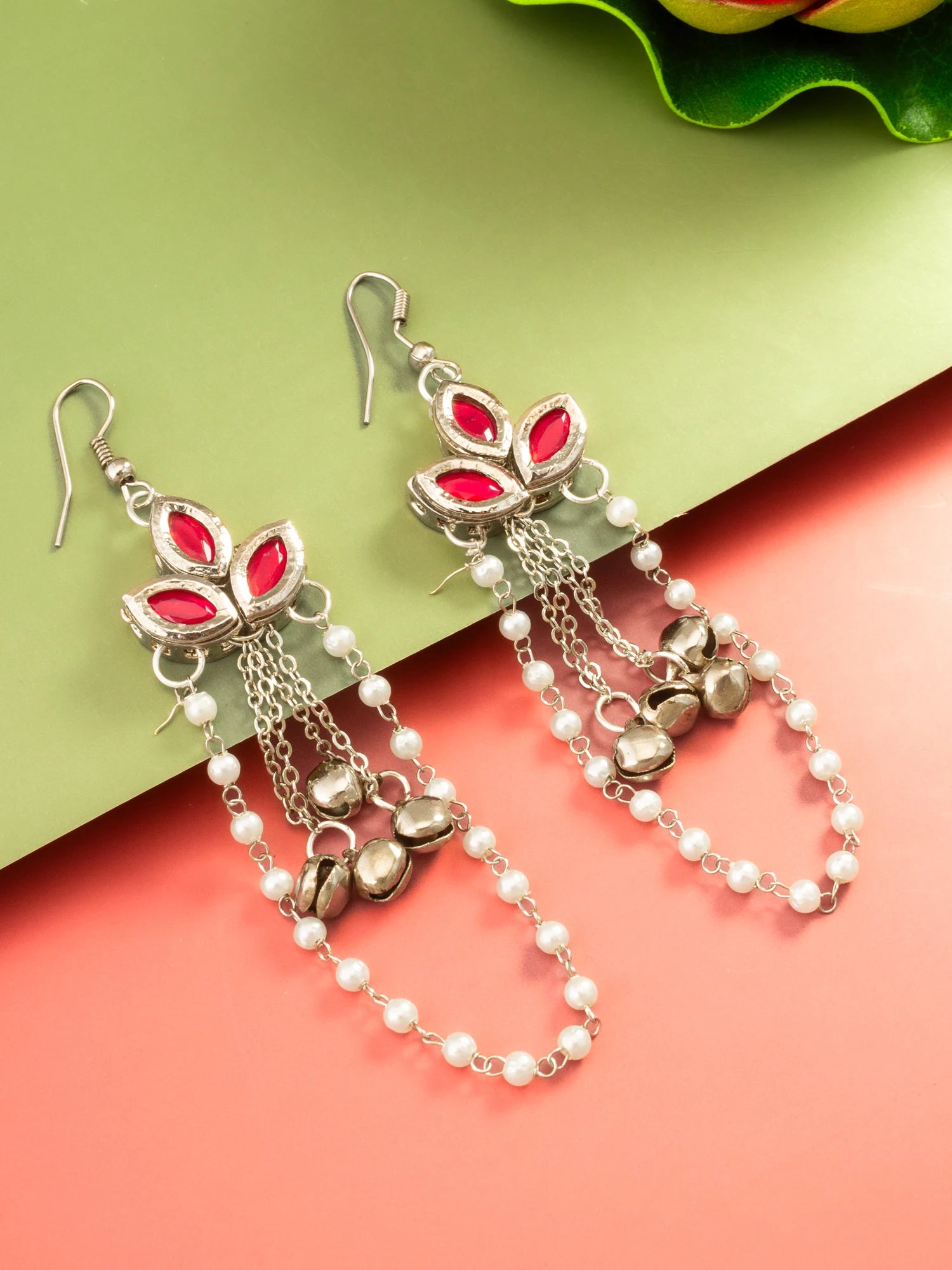 A pair of silver-toned dangle earrings with red gemstone-like accents, white pearl beads, and small metallic bells, displayed on a split green and coral background.