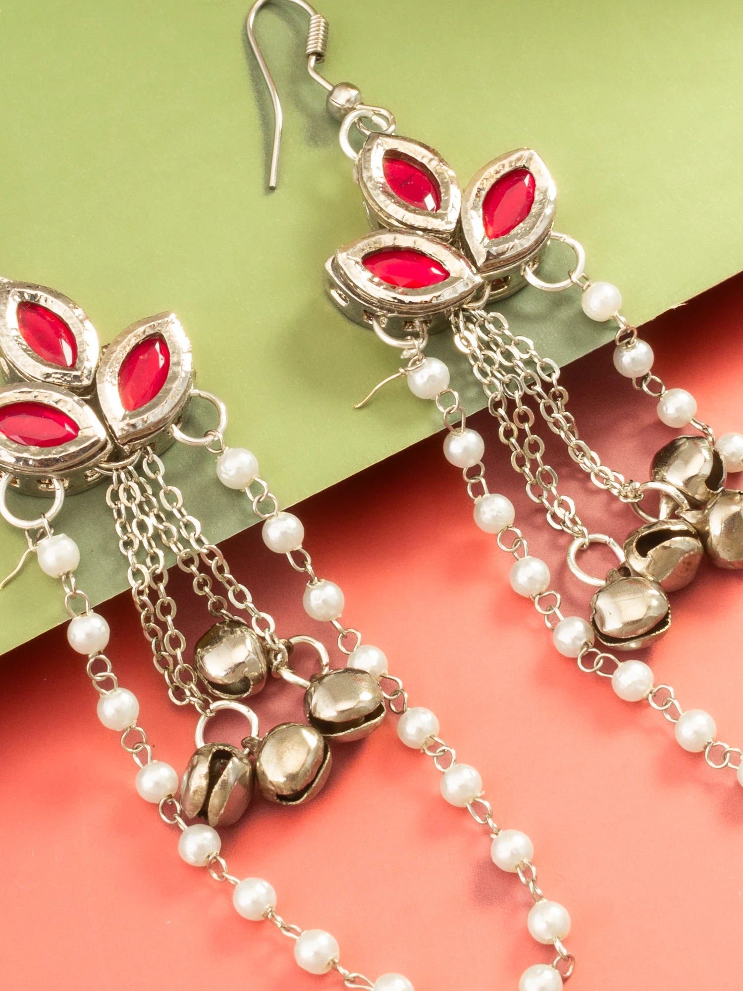 Silver-toned dangle earrings with red teardrop-shaped stones, white pearl beads, and small metallic bells, shown on a split green and coral background.