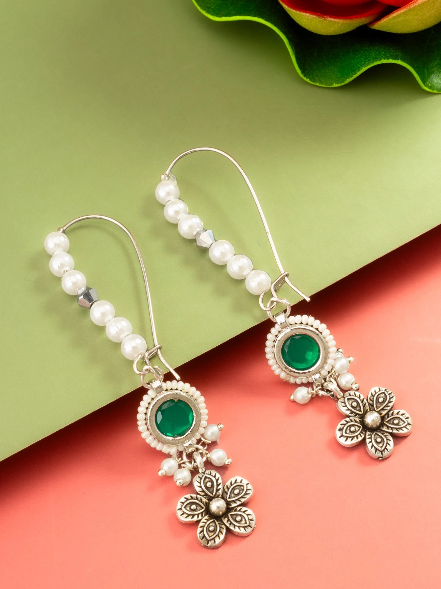 A pair of silver-toned dangle earrings with white pearl beads, oval green stone centers, and dangling floral charms, displayed on a split green and coral background.
