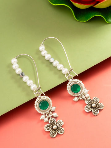 A pair of silver-toned dangle earrings with white pearl beads, oval green stone centers, and dangling floral charms, displayed on a split green and coral background.