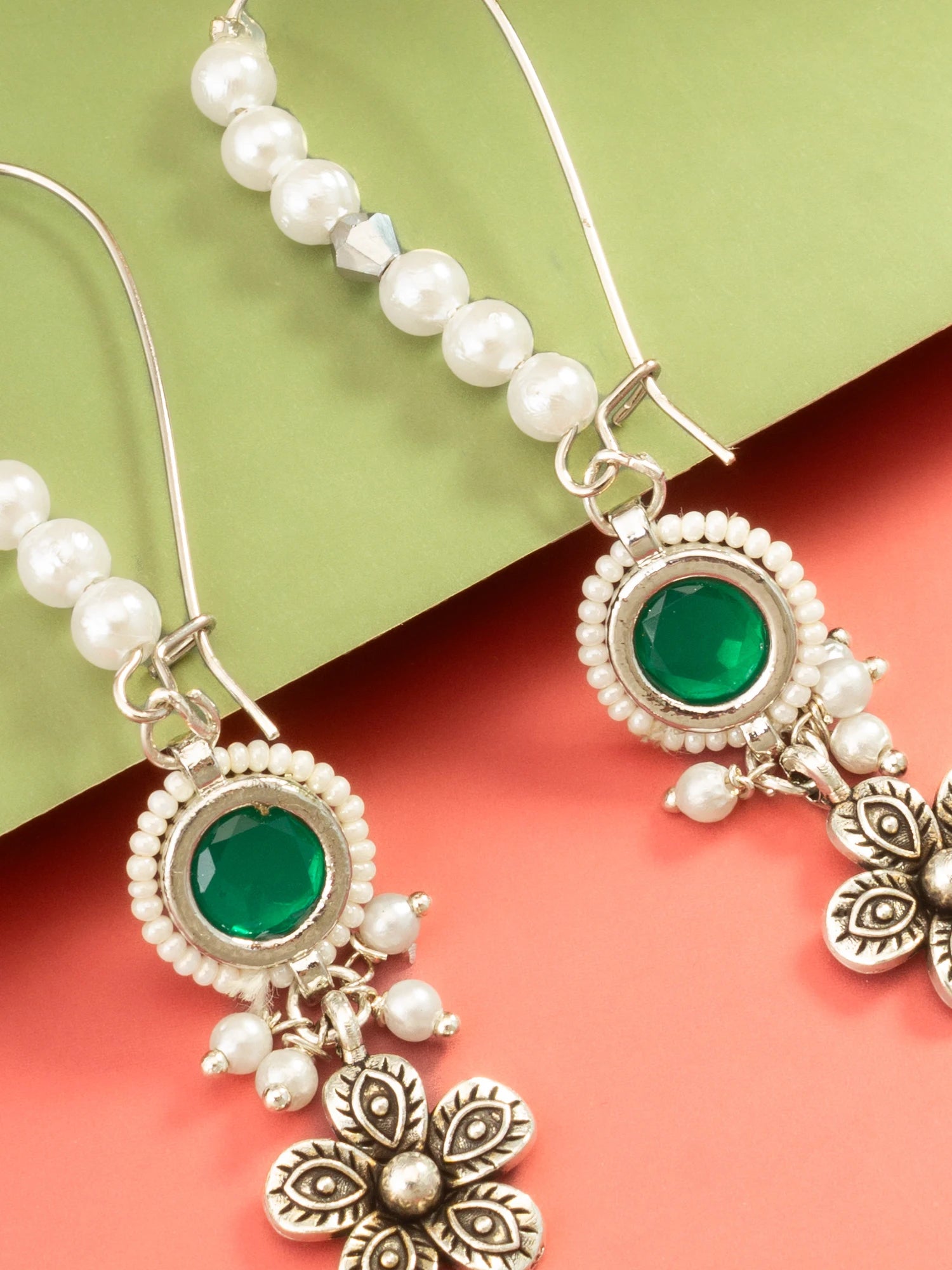 Silver-toned dangle earrings with white pearl beads, round green gemstones, and ornate floral charms, shown on a split green and coral background.