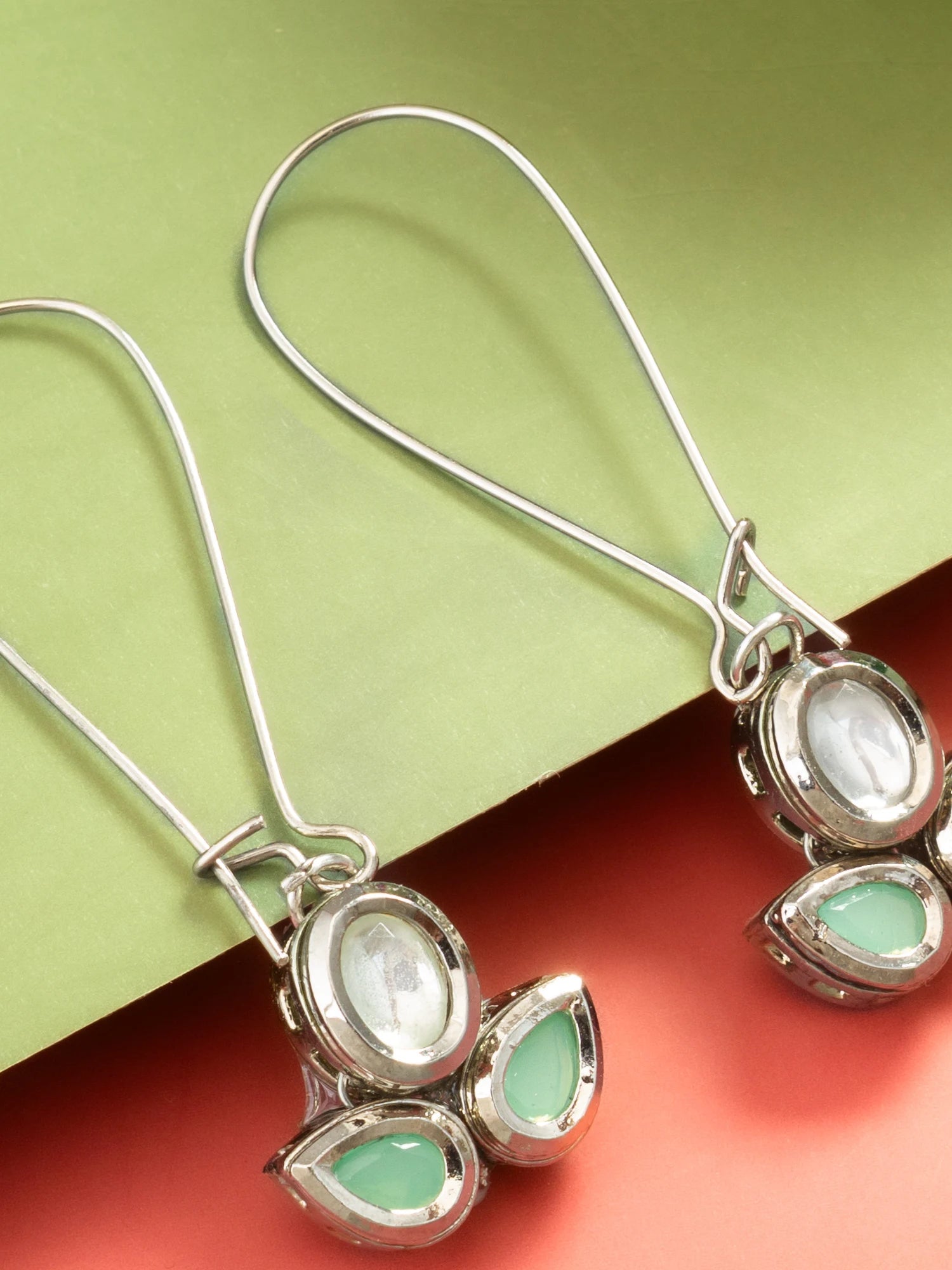 A pair of silver drop earrings with oval and teardrop-shaped light green gemstones, displayed on a split green and coral background.