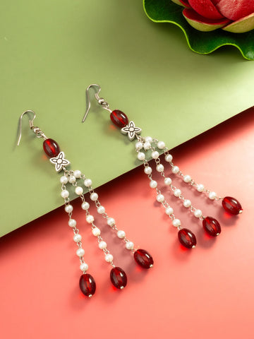 A pair of silver-toned dangle earrings with red faceted beads and white pearl-like beads, displayed on a split green and coral background.
