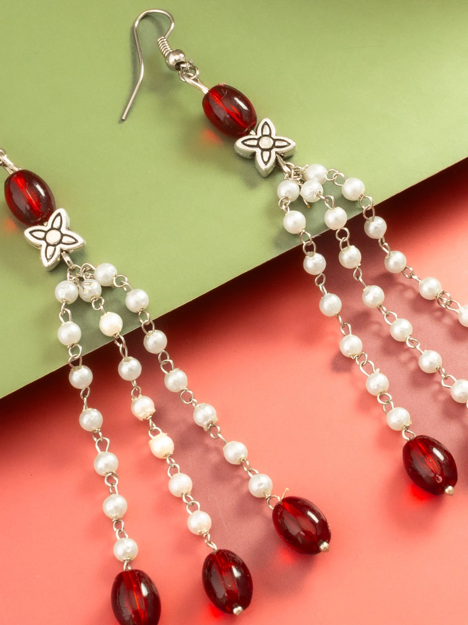 A pair of dangling earrings with silver-tone metal hooks and flower-shaped charms, accented with white pearl beads and deep red faceted beads, shown on a split green and coral background.