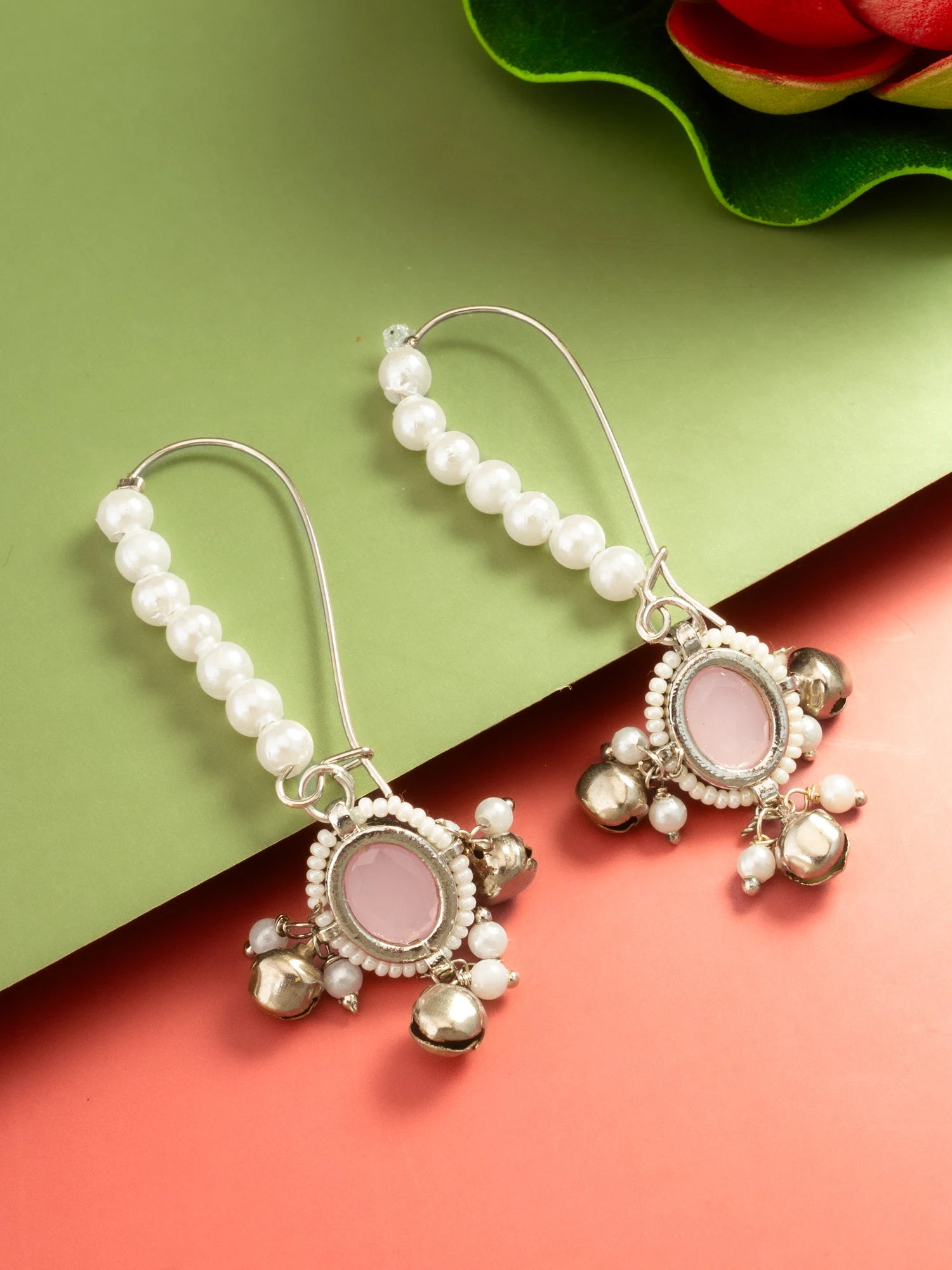 A pair of silver-toned dangle earrings with white pearl bead strands, oval pink stone centers, and dangling metallic bell charms, arranged on a green and coral background.