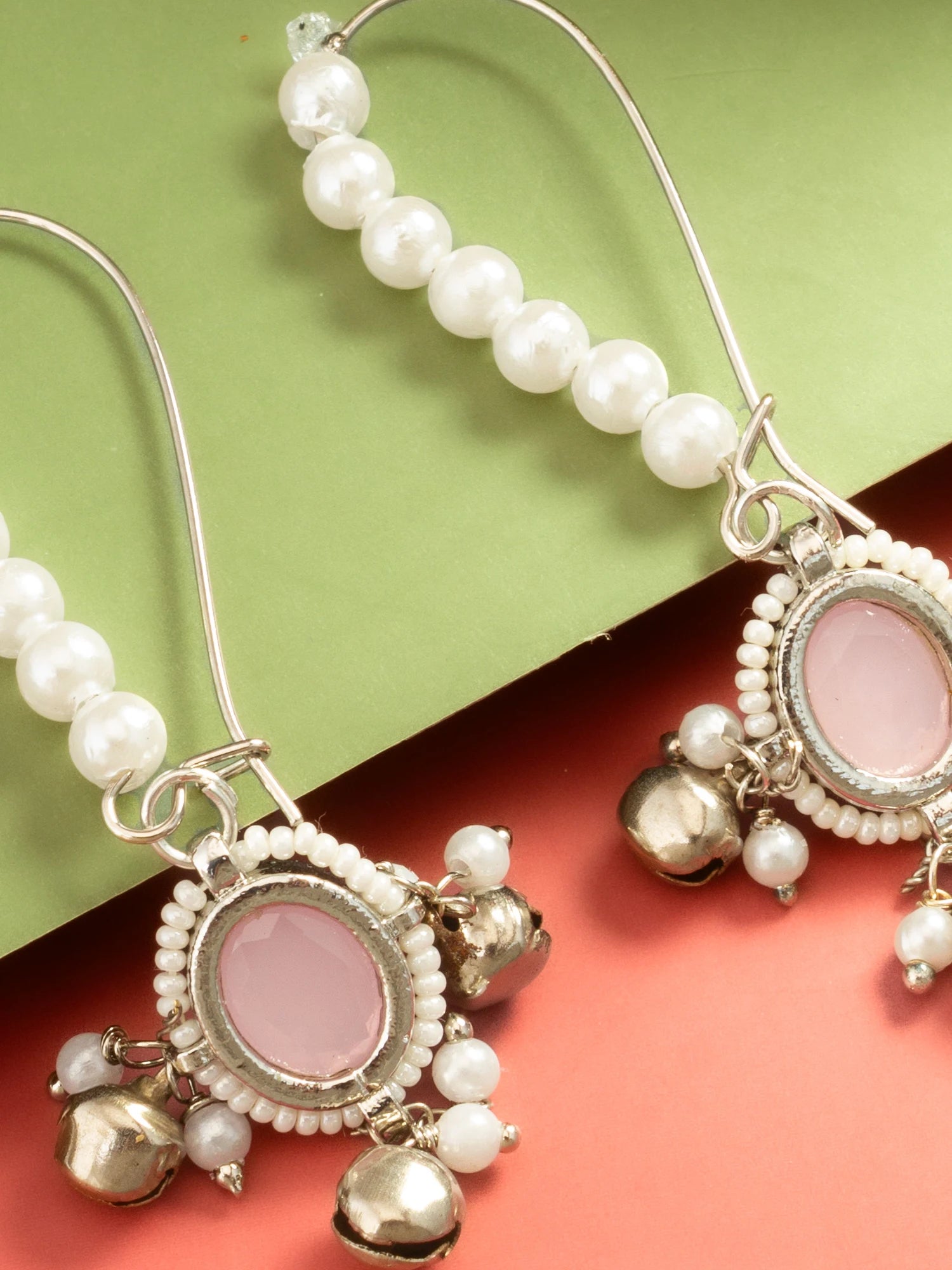 A pair of silver-toned dangle earrings with white pearl bead strands, oval pink stone centers, and small dangling bell charms, shown on a split green and coral background.