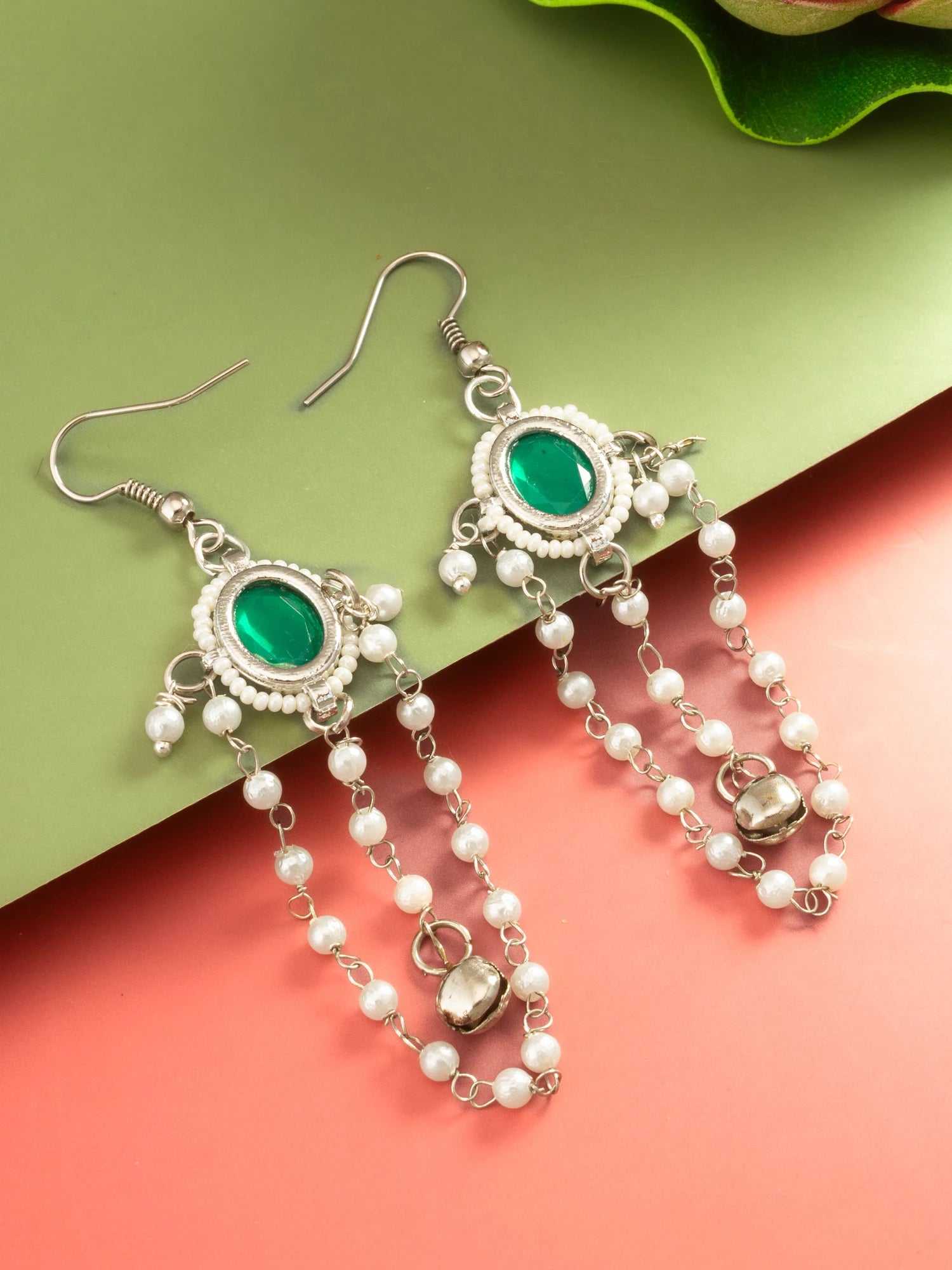 A pair of silver-toned dangle earrings with oval green stones and white pearl chains, displayed on a split green and coral background.