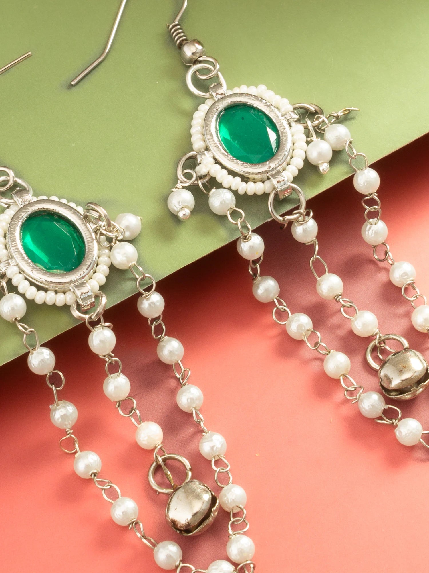A pair of silver-toned traditional Indian earrings with oval green stones and dangling pearl bead chains, displayed on a split green and coral background.