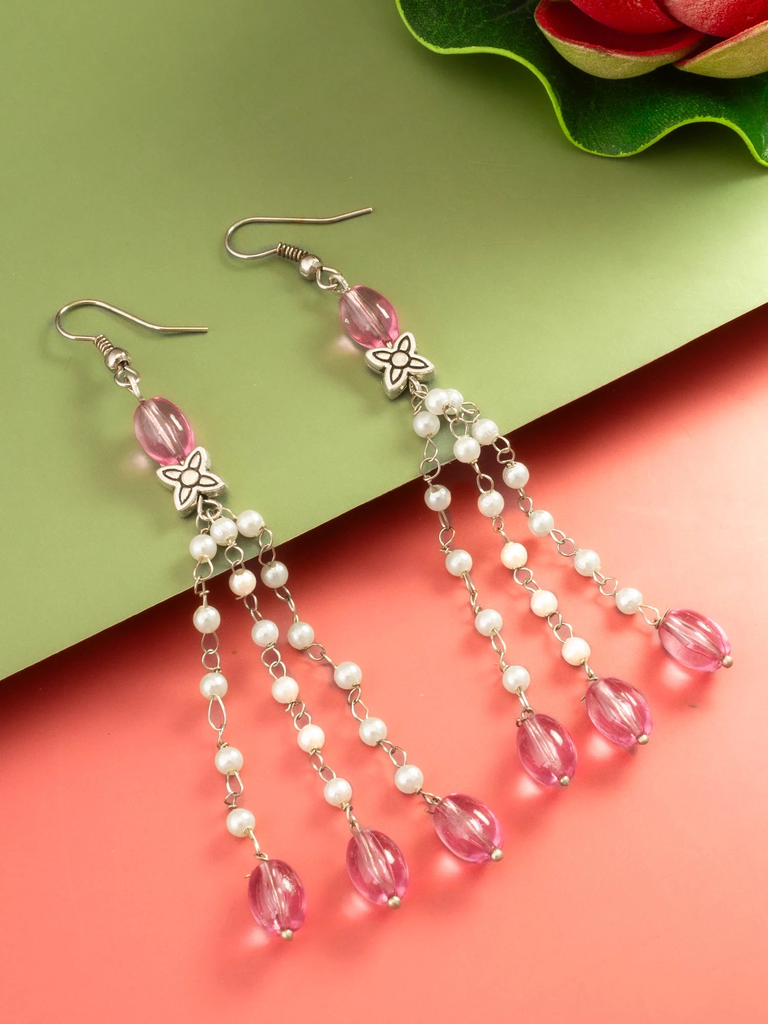 A pair of silver-toned dangle earrings with pink faceted beads, white pearl-like beads, and silver flower charms, displayed on a split green and coral background.