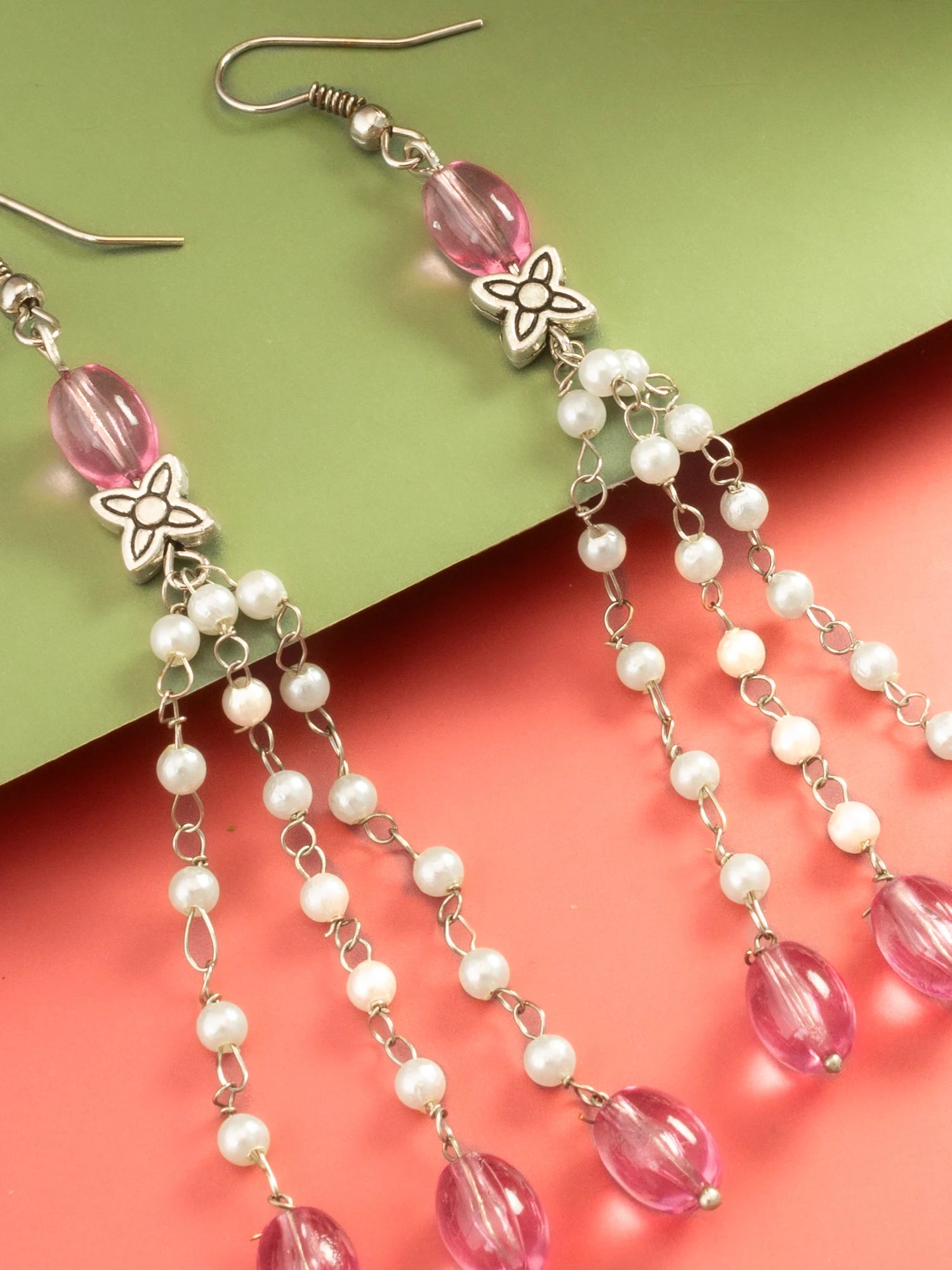 Pink and white beaded dangle earrings with silver-tone hooks and floral charms, displayed on a split green and coral background.