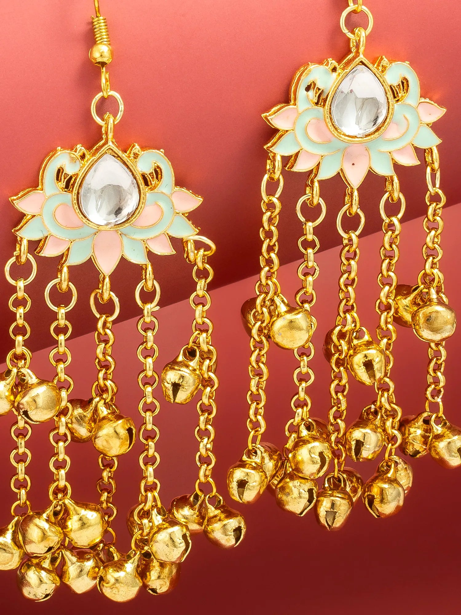 Gold-toned dangle earrings with lotus-shaped floral tops, pale mint green and pink enamel details, clear teardrop stones, and multiple dangling bell chains on a pink background.
