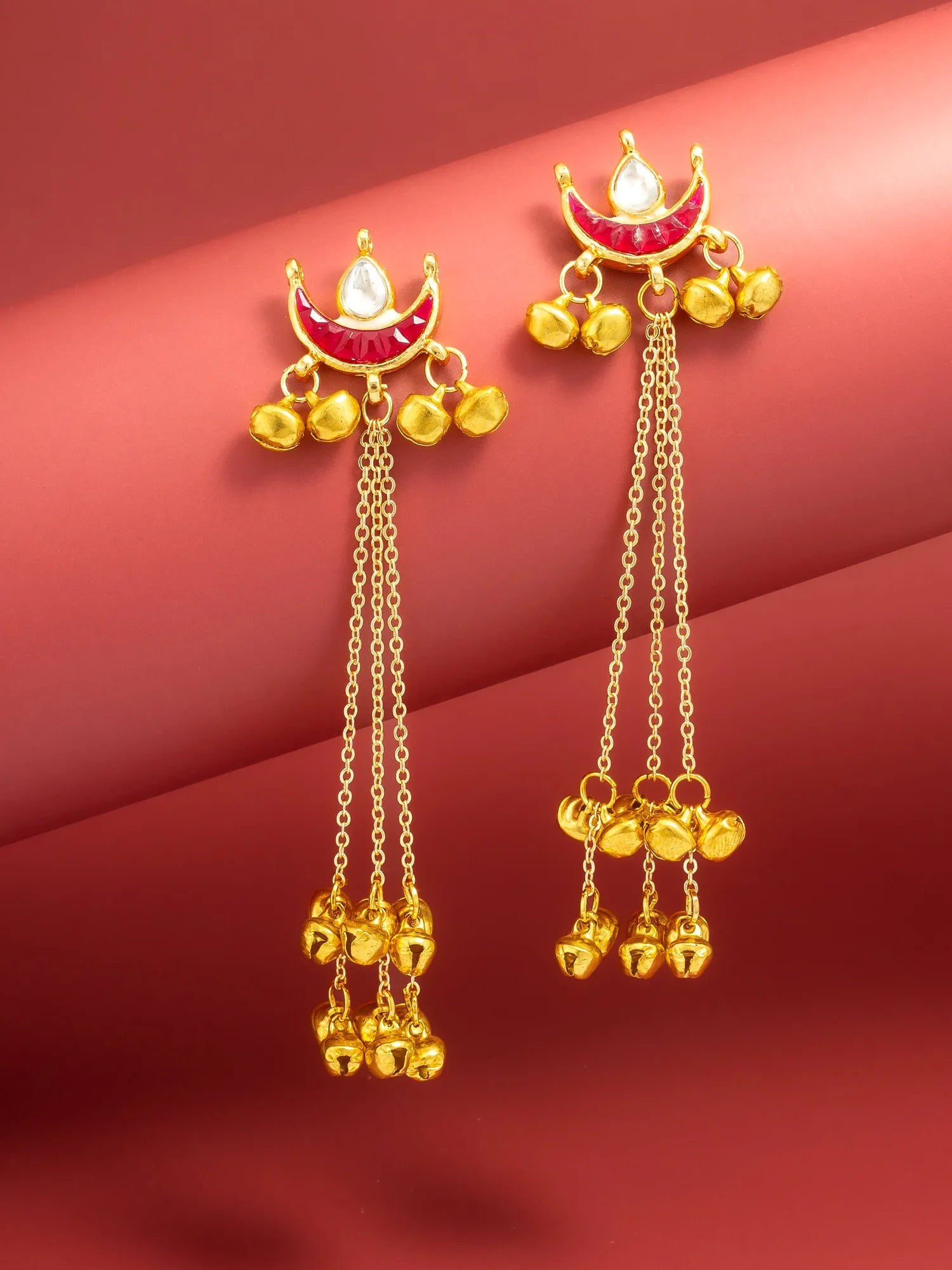A pair of gold-toned traditional Indian dangle earrings with red enamel crescent moon details, white stones, and dangling bell charms, displayed on a curved red background.