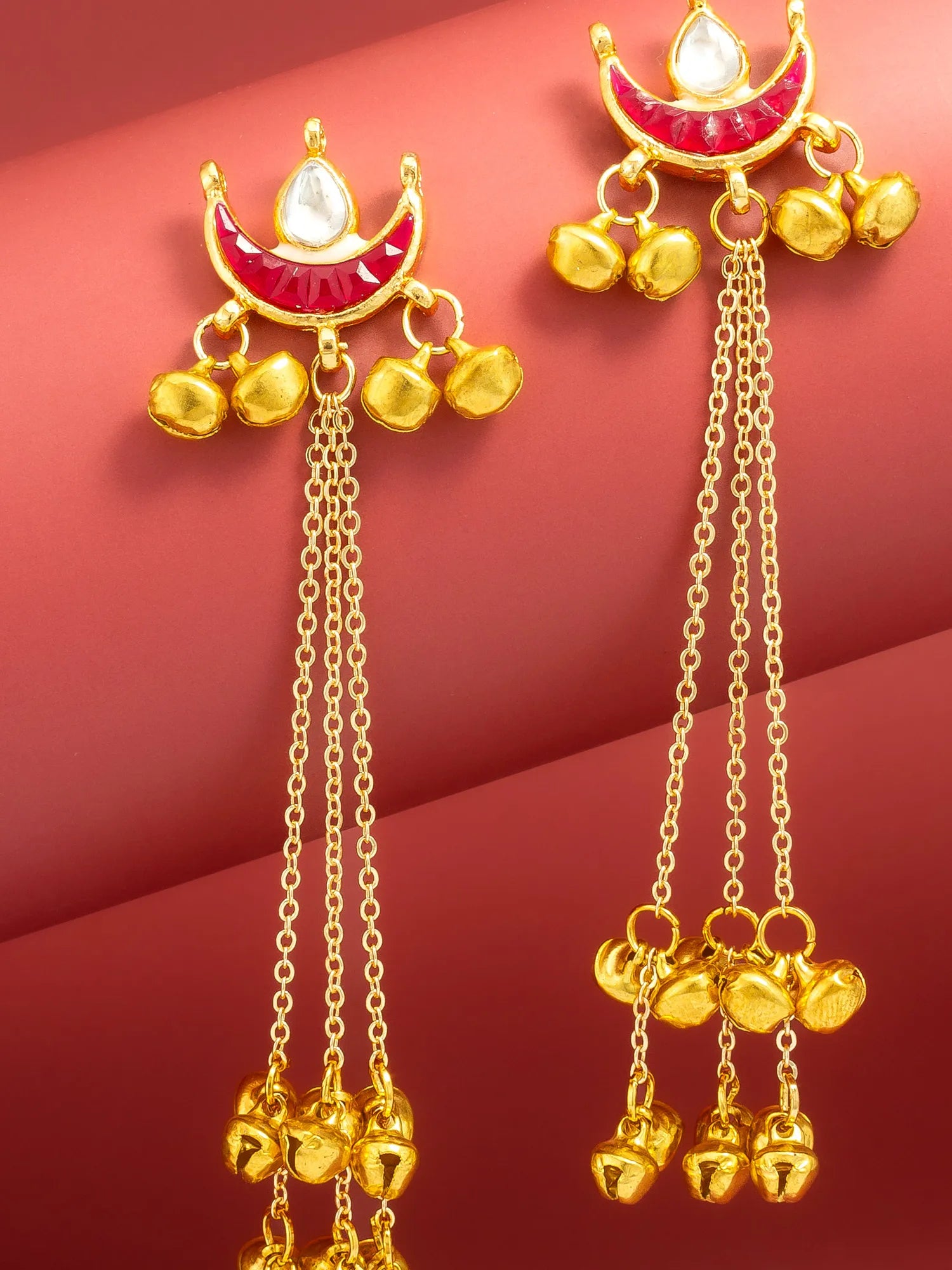 Gold-toned traditional Indian dangle earrings with red enamel crescent motifs, white stone accents, and dangling bell charms, shown on a red background.