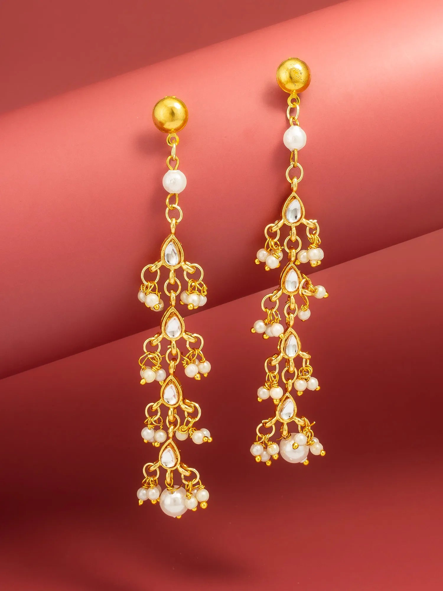 A pair of gold dangle earrings with white pearl-like beads and teardrop crystal accents, displayed against a warm pinkish-red gradient background.