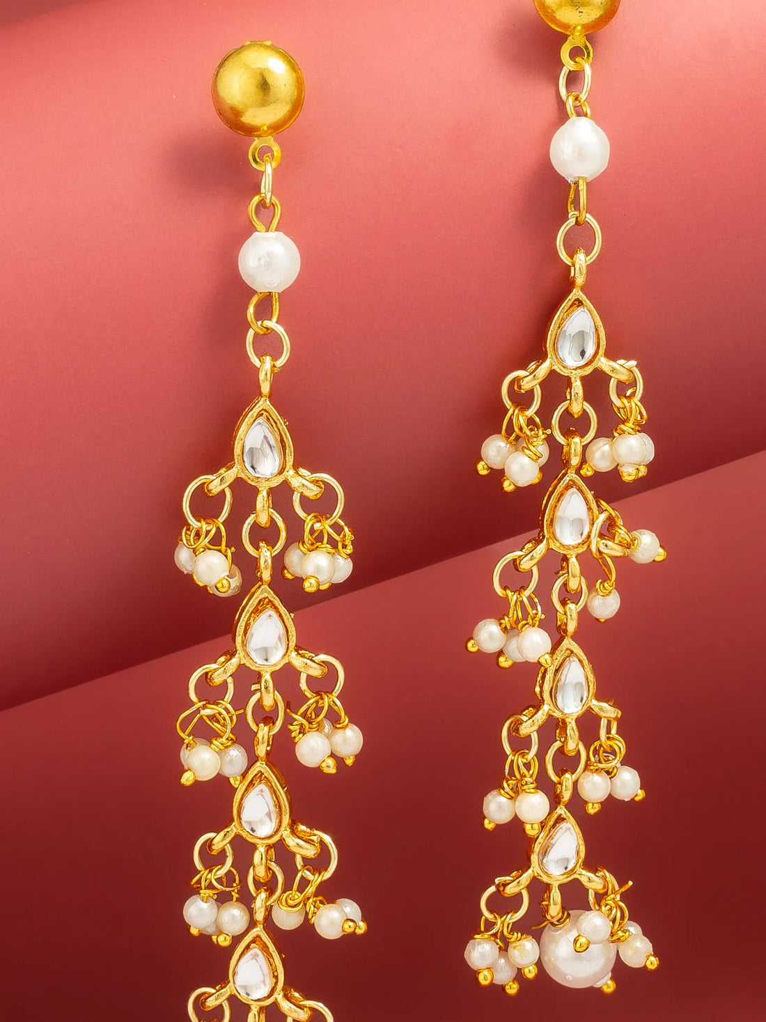 A pair of gold dangle earrings with white pearl-like beads and teardrop crystal accents, displayed against a warm pinkish-red gradient background.
