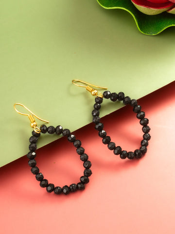 Black faceted bead hoop earrings with gold fishhook ear wires, displayed on a split green and pink background with a decorative green leaf in the corner.
