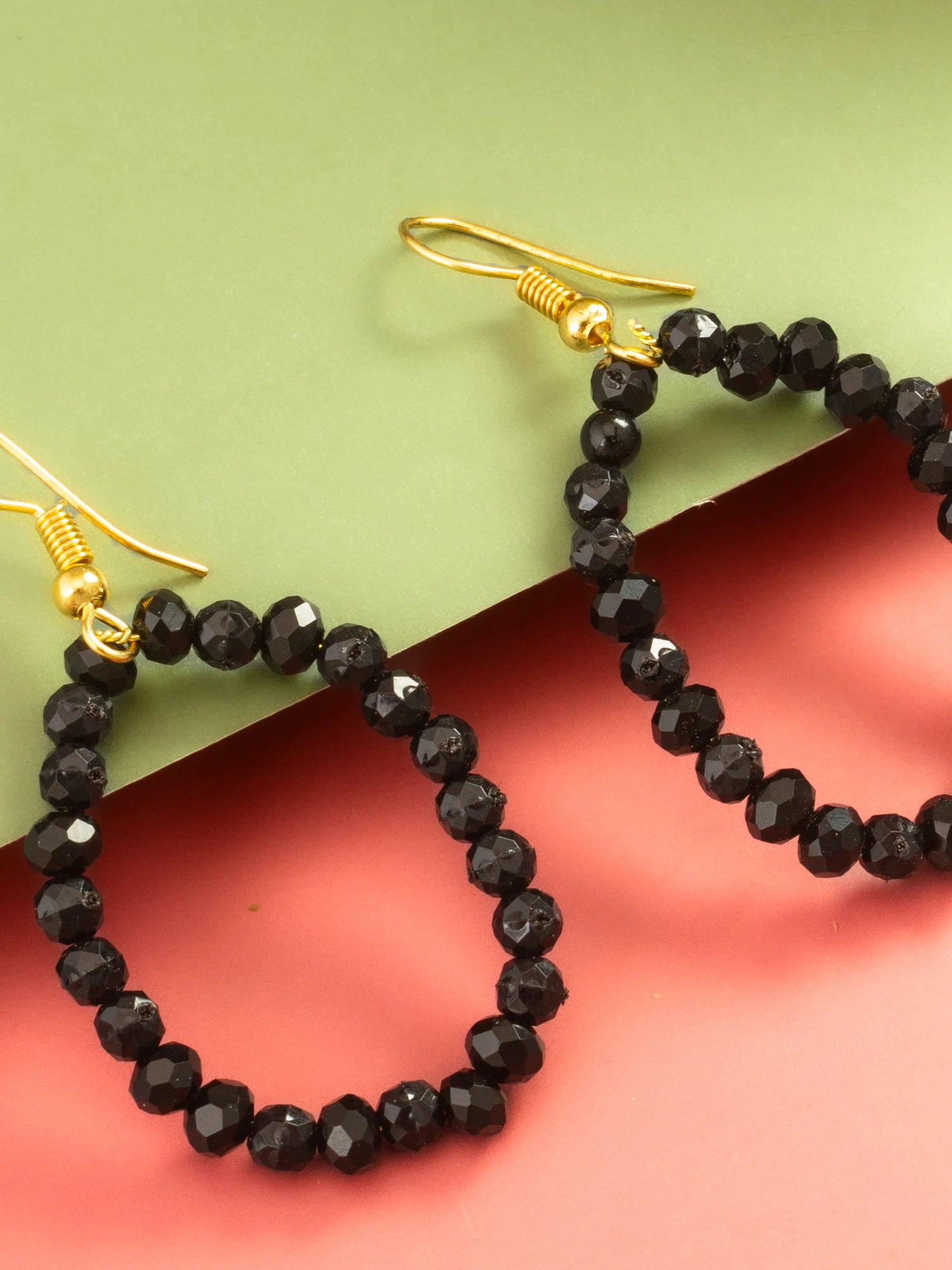 Black faceted bead hoop earrings with gold fishhook ear wires, displayed on a split green and coral pink background.