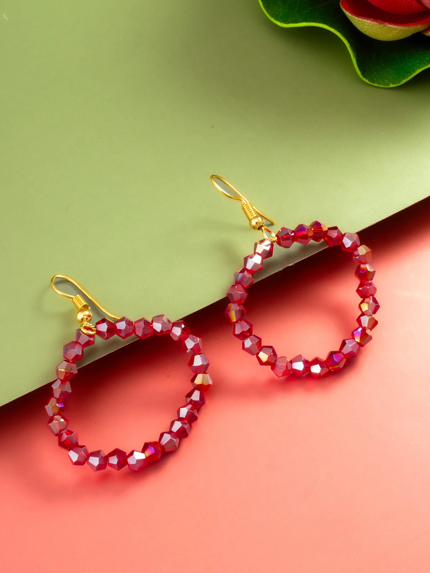 A pair of gold-tone hoop earrings with faceted deep pink beads, displayed on a split green and coral background.