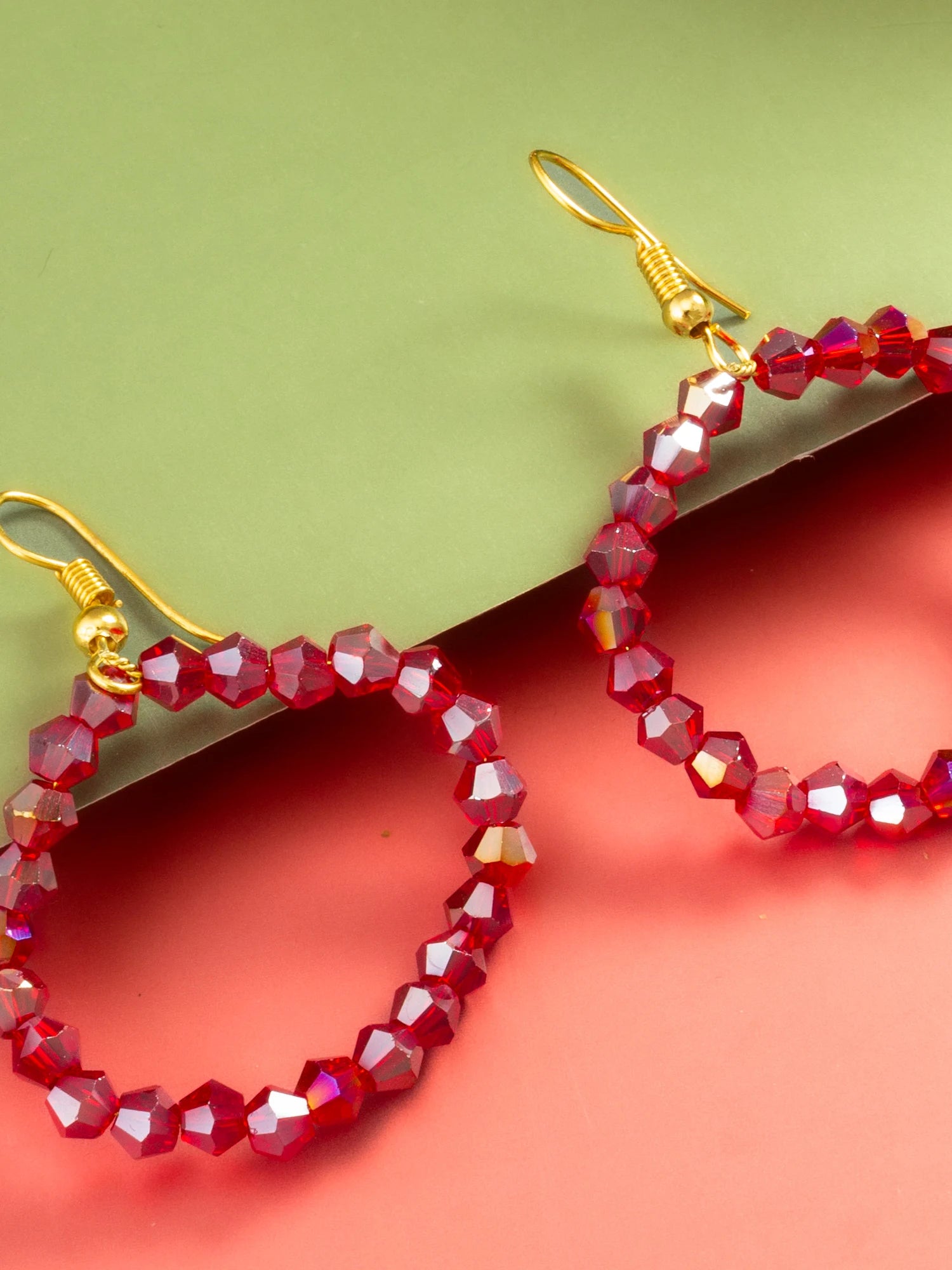 Red faceted bead hoop earrings with gold fishhook ear wires, displayed on a split green and coral pink background.
