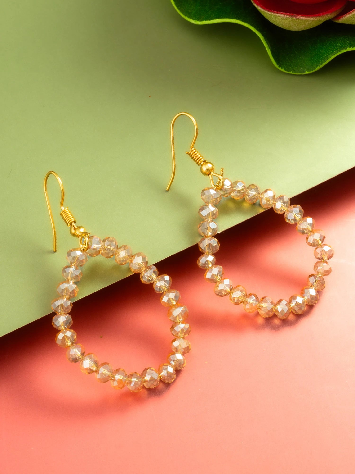 A pair of gold-tone drop earrings with faceted pearl-like beads, displayed on a split green and coral pink background.